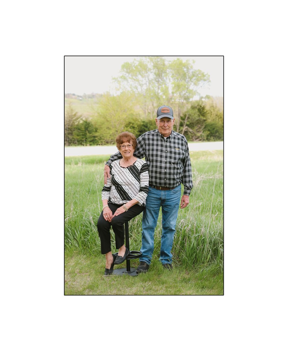 Older couple posing outdoors in a green field, woman seated and man standing behind her, trees in background.