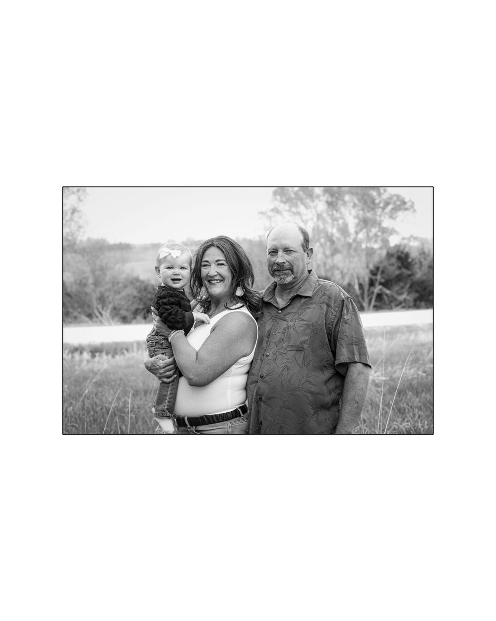 Black and white portrait of a couple smiling while holding a young baby in a grassy outdoor setting.