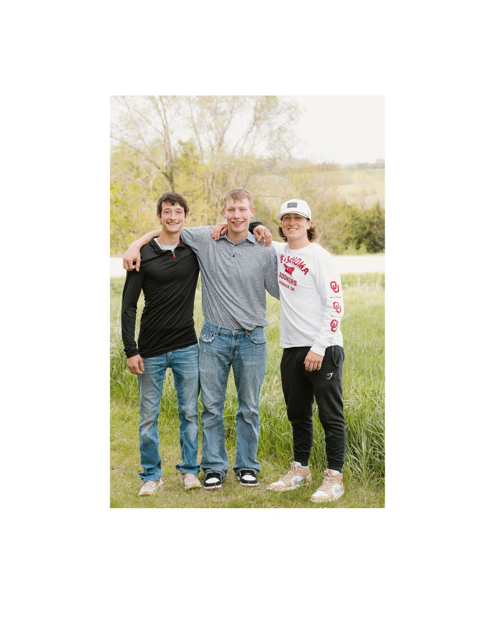 Three young adults smiling together in a green outdoor field on a sunny day.