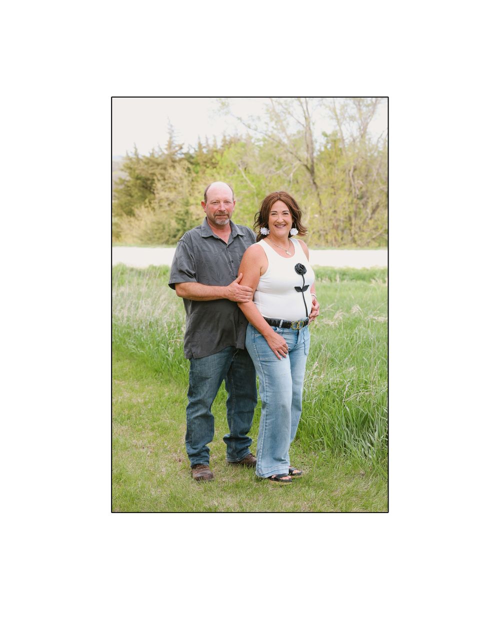 Middle-aged couple smiling and embracing outdoors in a lush green field with trees and water behind them.