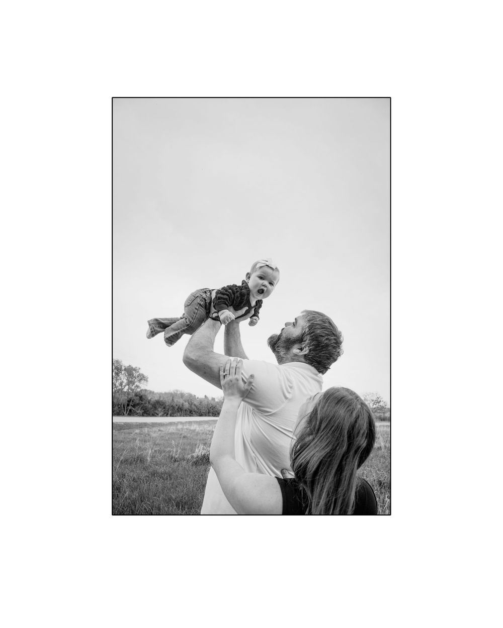 A father joyfully lifts his laughing baby in the air while the mother watches in a grassy field.