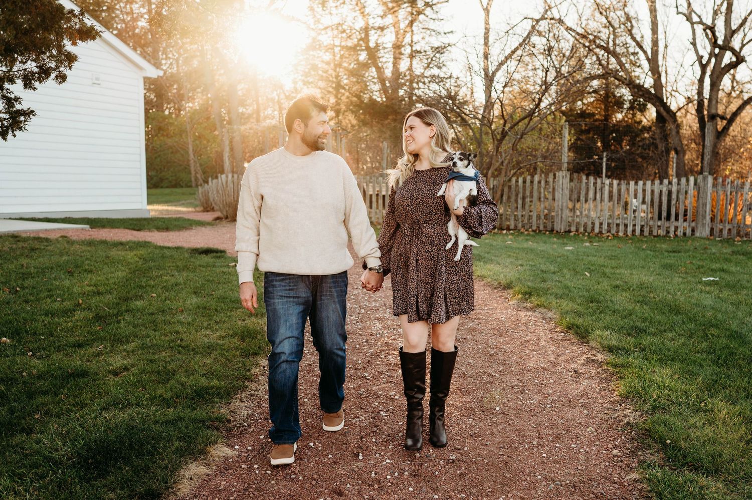 Couple holding hands walking on a dirt path at golden hour with a white building and wooden fence in background.