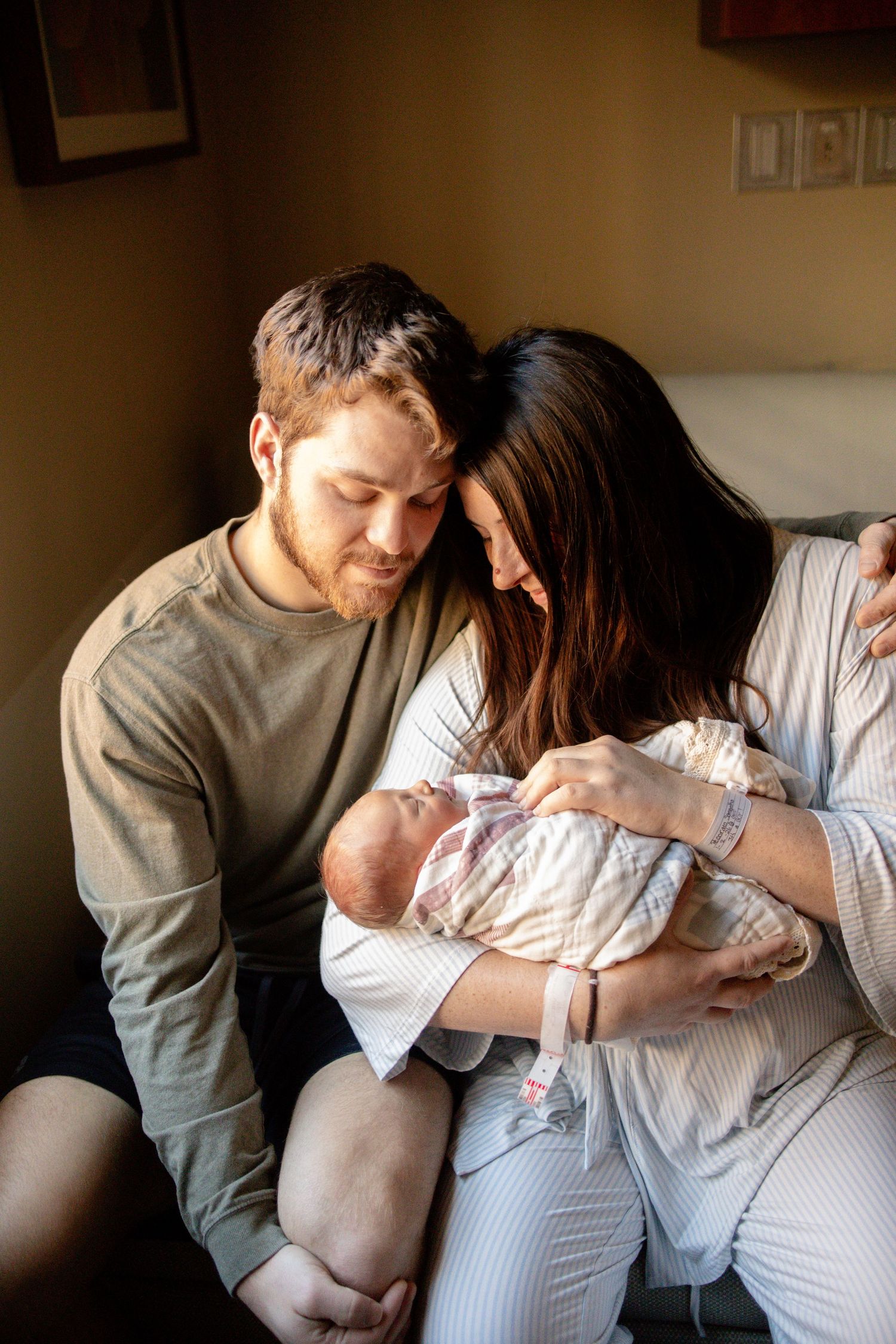 New parents lovingly gaze at their newborn baby in a warm hospital room, mom in a striped robe cradling the infant.