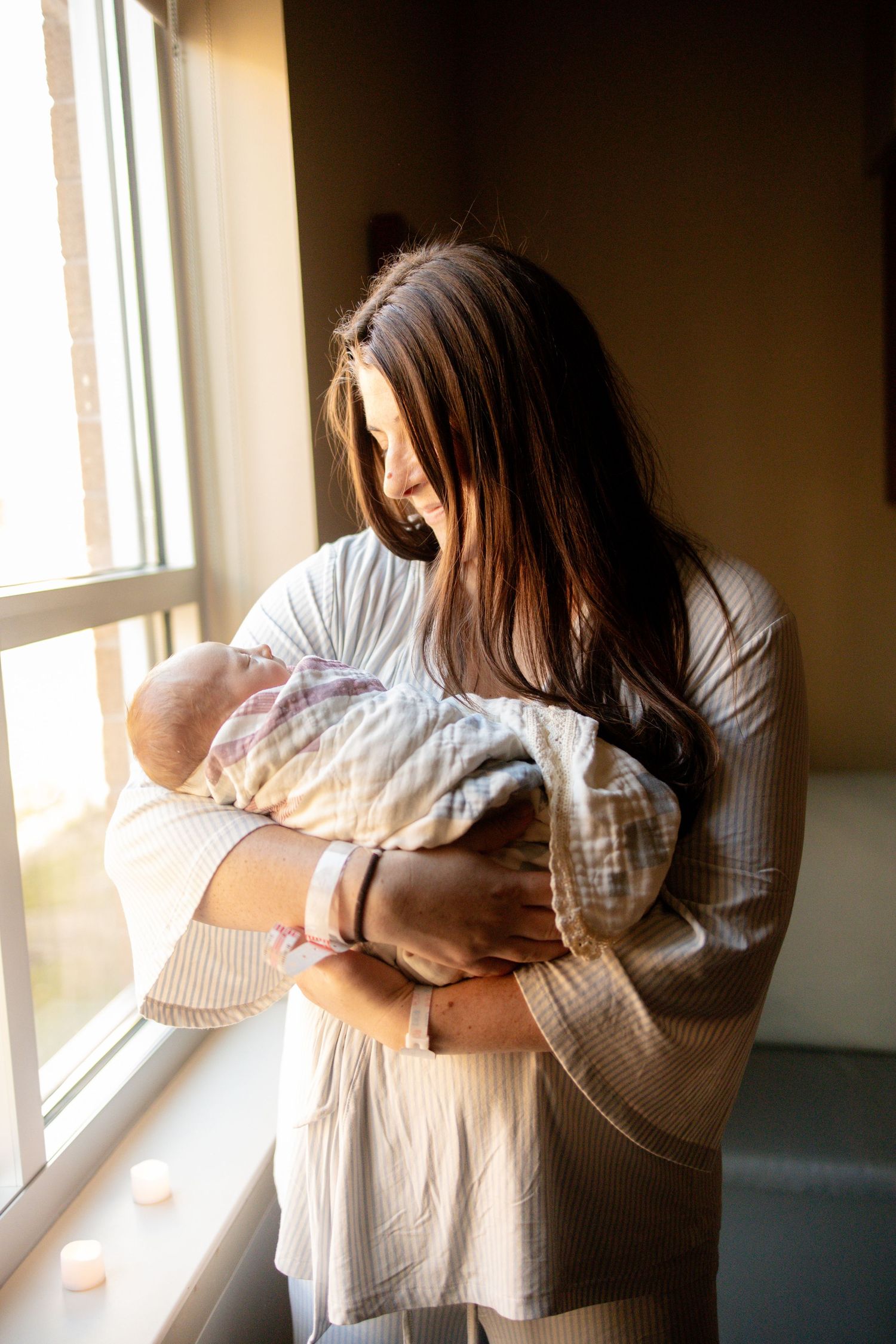 A mother with long brown hair gently holds her newborn baby near a sunlit window indoors.