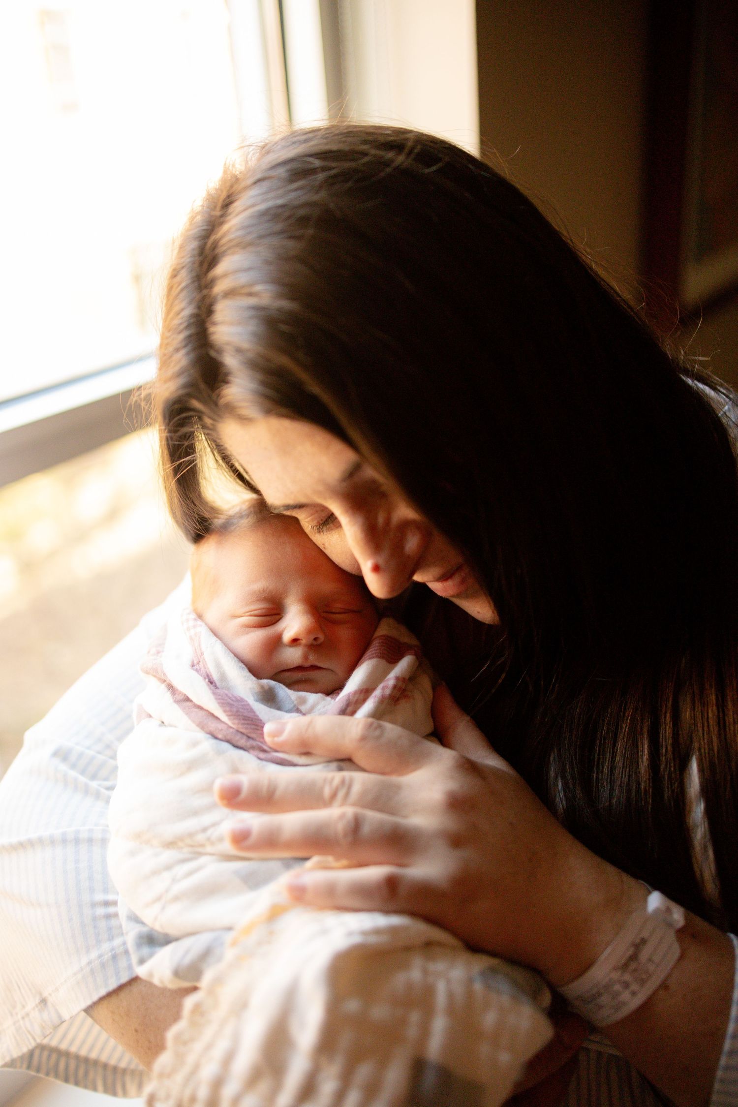 A dark-haired mother tenderly holds her swaddled newborn baby close to her face near a bright window.