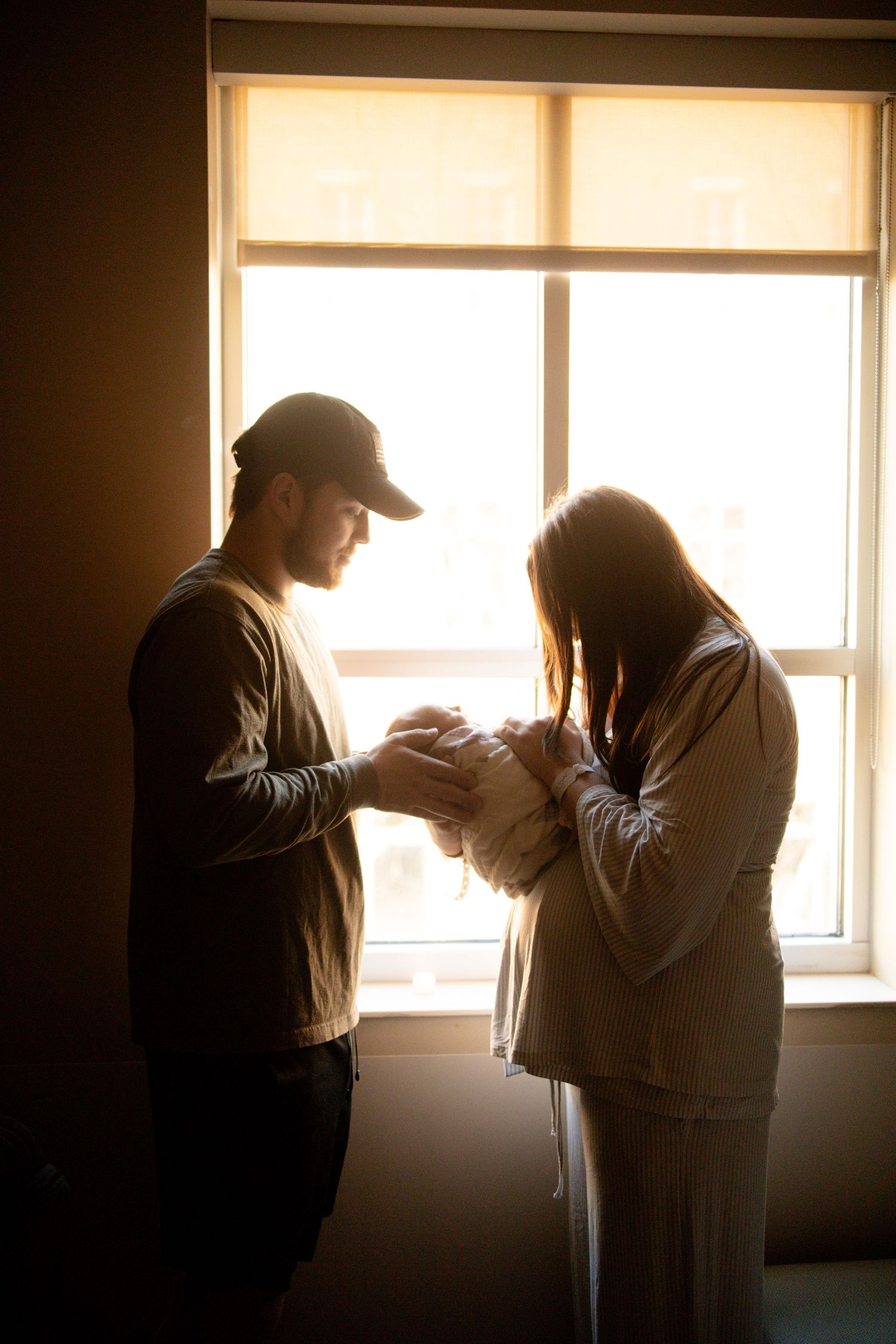New parents standing by a sunlit window, tenderly holding their newborn baby in a warm, intimate hospital room.