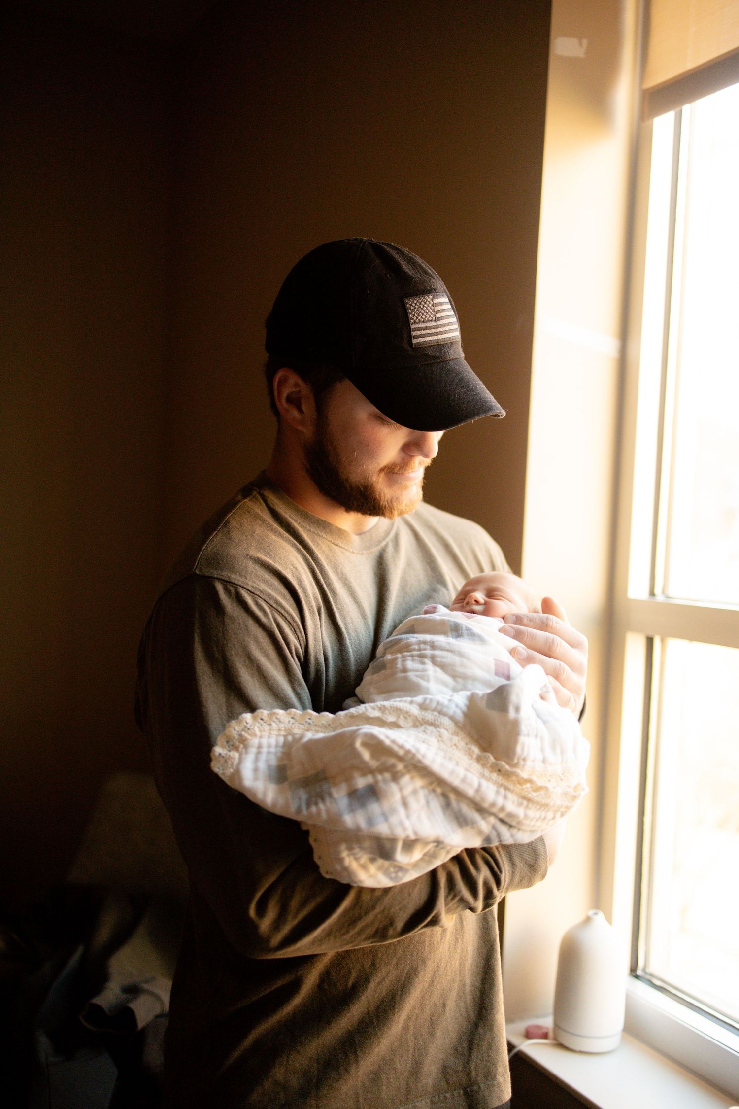 A bearded man in a black cap tenderly holds a newborn baby wrapped in white muslin near a sunlit window.