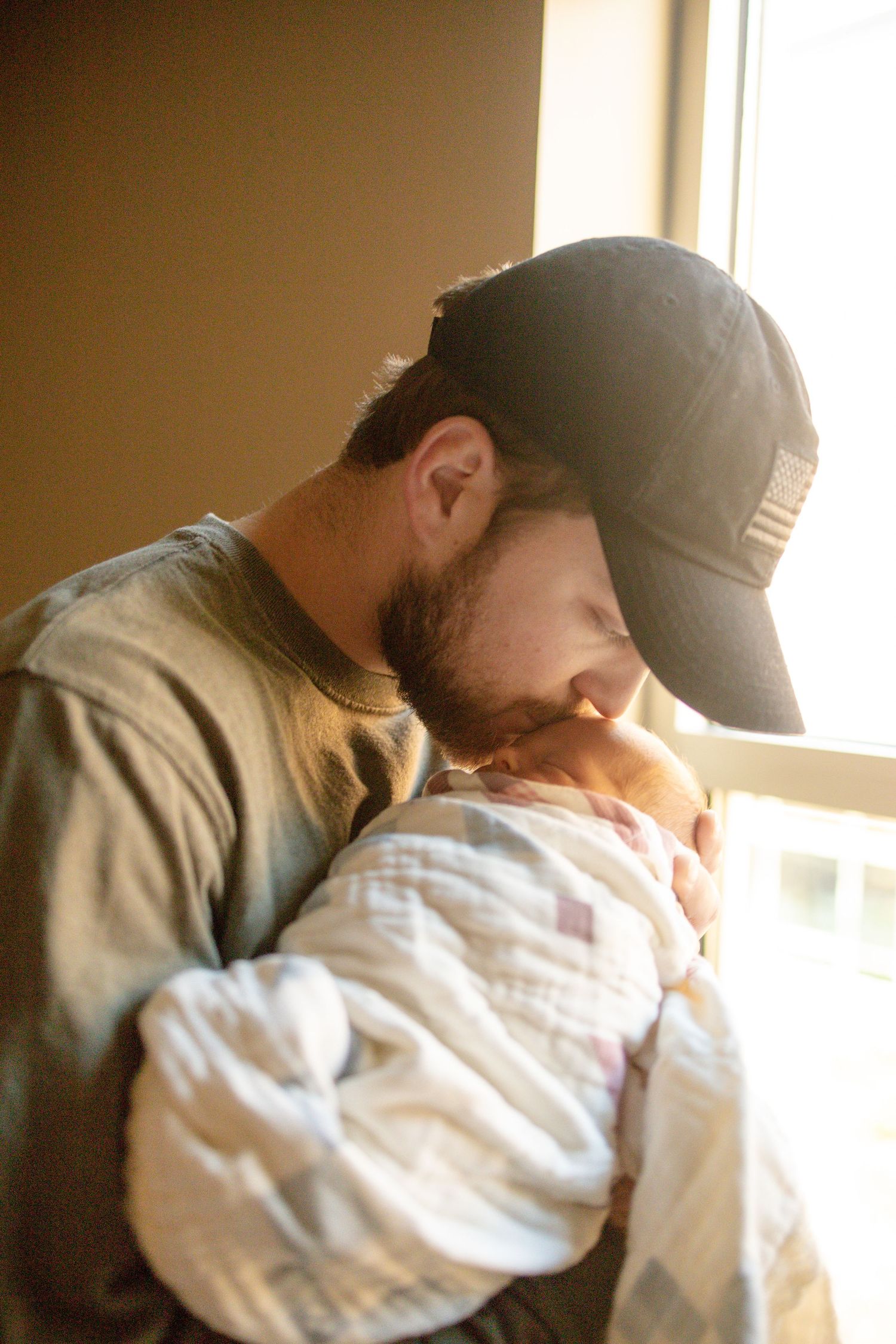 A bearded father in a baseball cap tenderly kisses his newborn baby wrapped in white swaddle near a sunlit window.