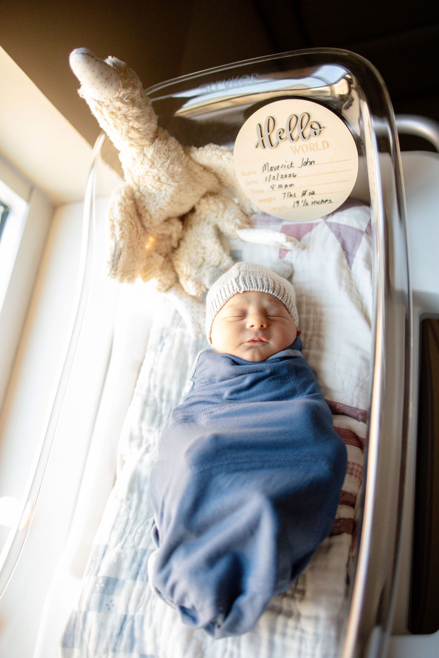 Newborn baby swaddled in blue blanket wearing white knit hat sleeping in hospital bassinet with stuffed bunny and Hello sign.