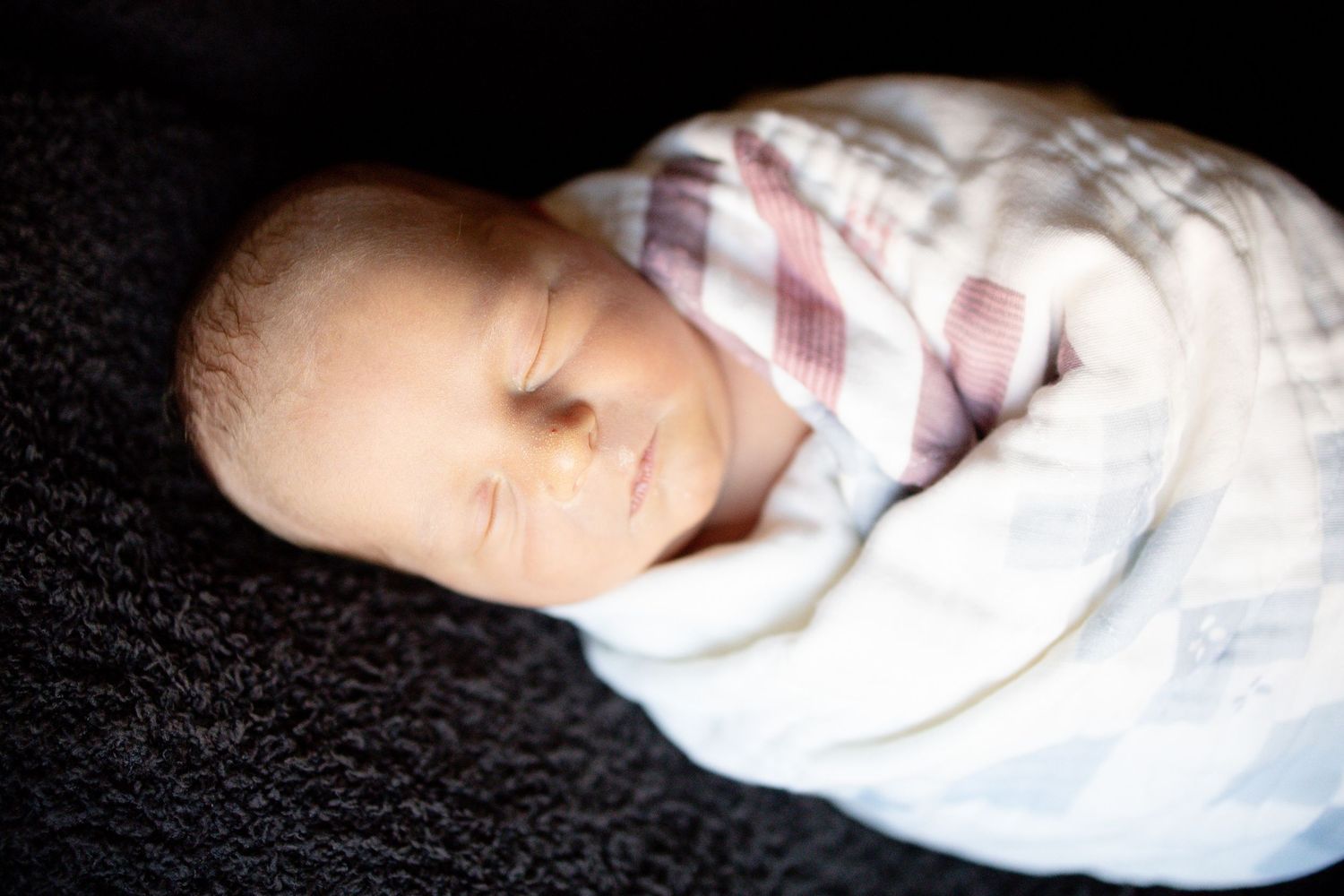 Peaceful newborn baby swaddled in a soft white and plaid blanket, sleeping on a dark fuzzy surface.