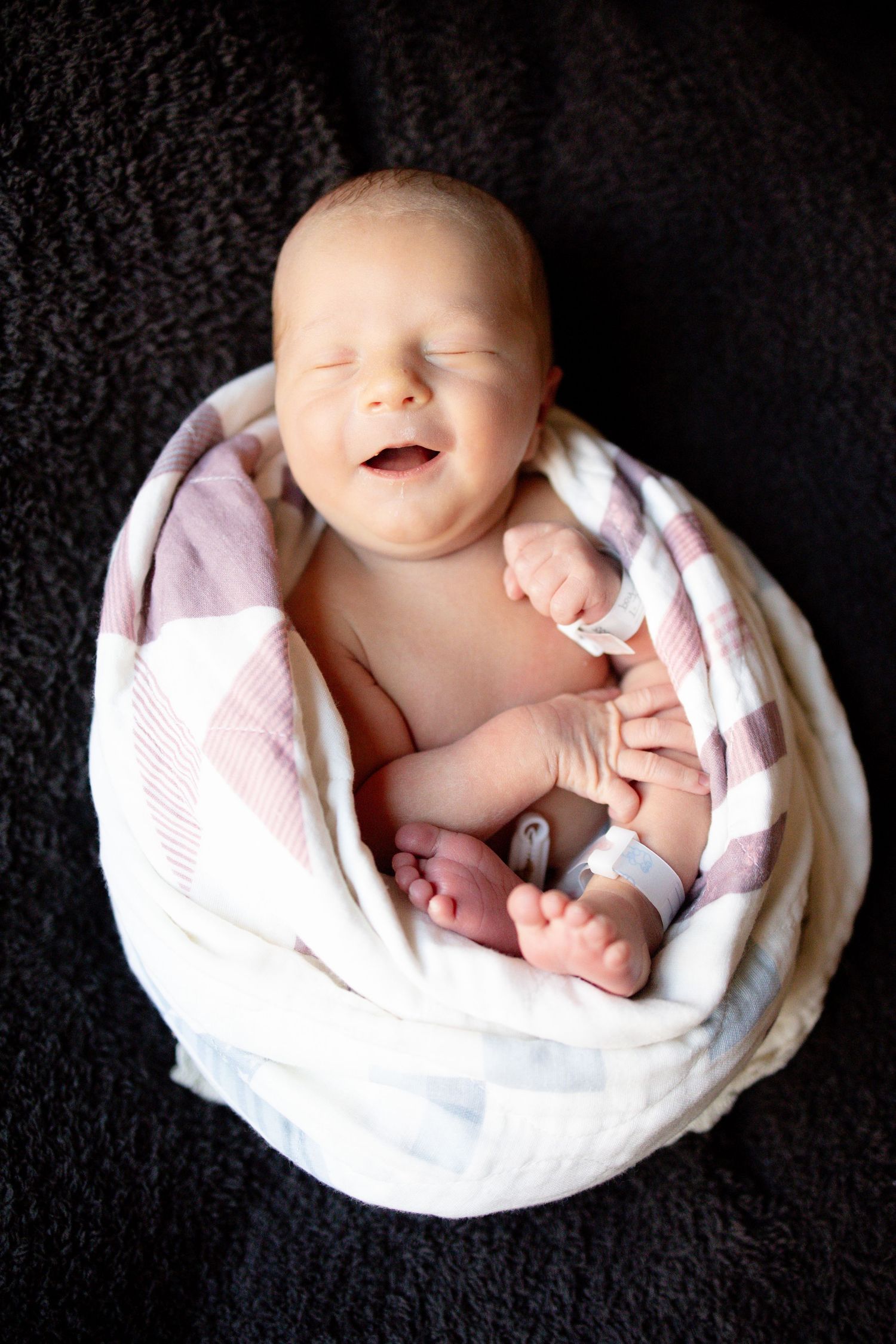 Smiling newborn baby wrapped in a pink and white blanket, lying on a dark background.