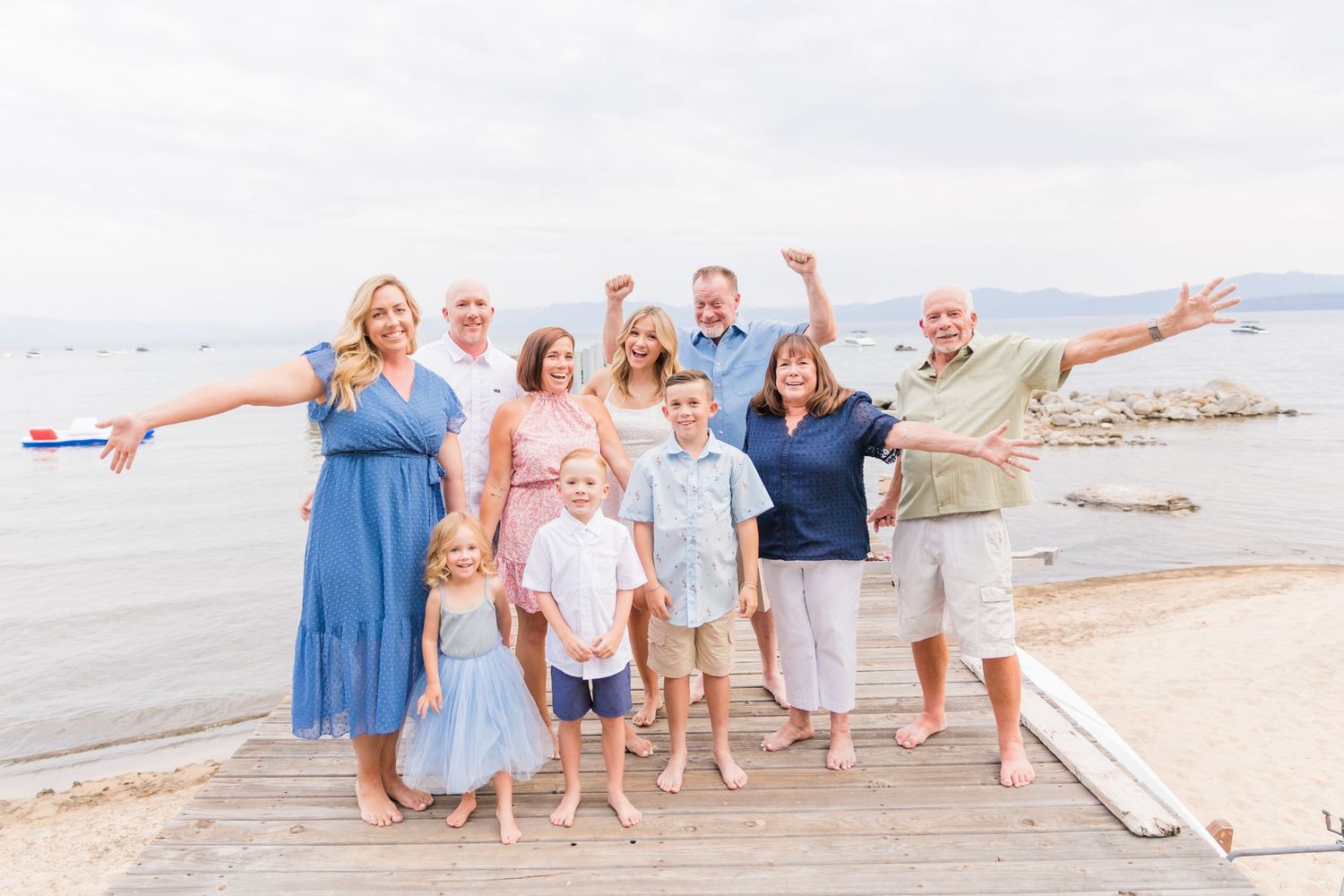 Large family group posing joyfully on a wooden dock by the beach, arms raised, on a cloudy day.