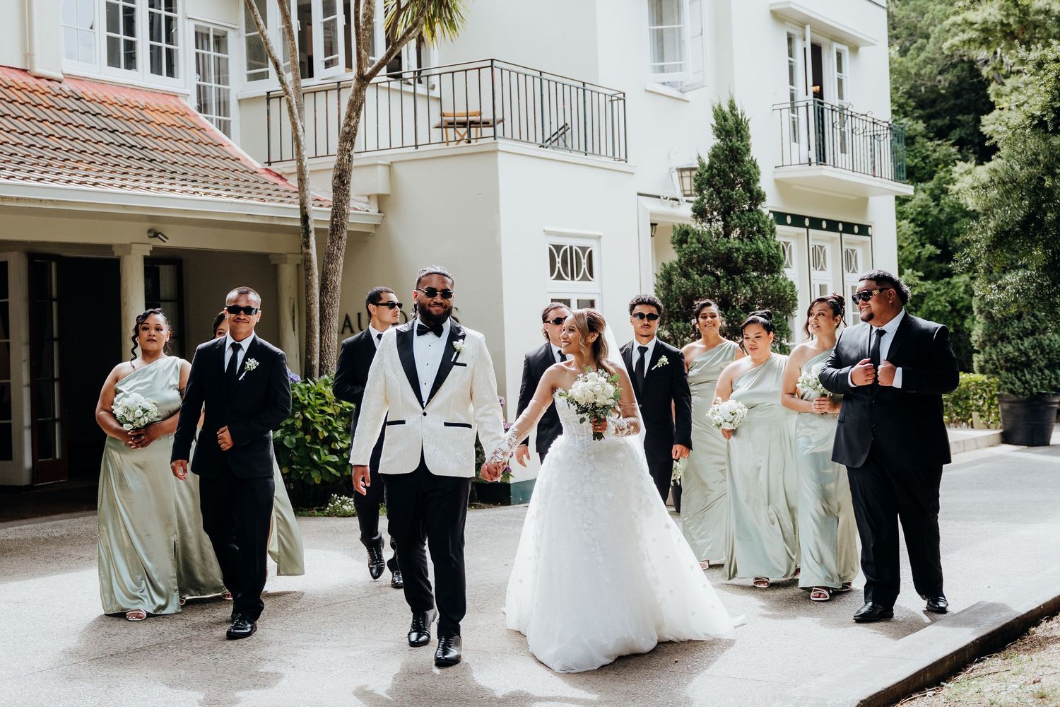Wedding party walks confidently outdoors, bride in white gown, groom in white tuxedo, bridesmaids in sage green dresses.