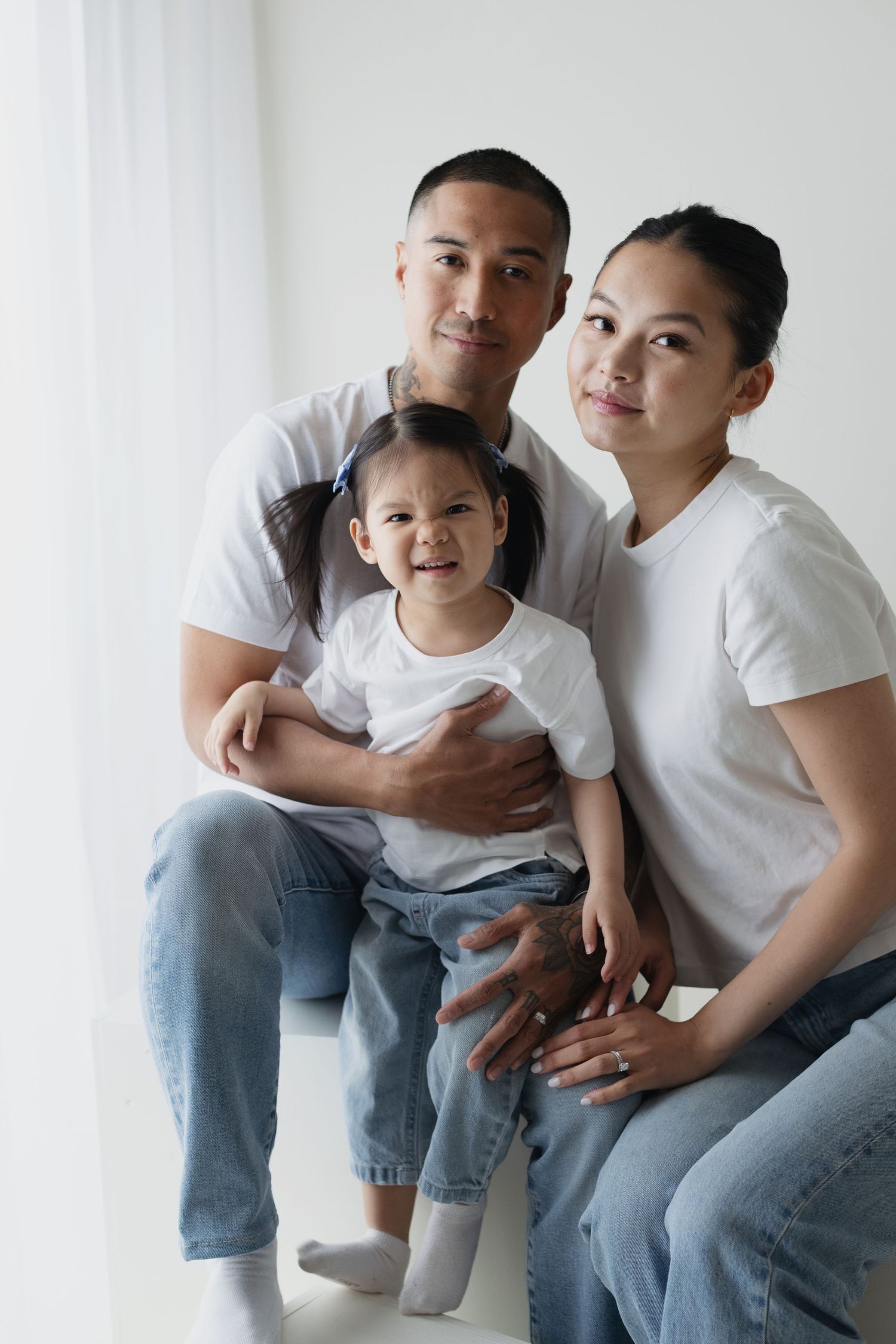 Happy Asian family with young daughter in white shirts and jeans smiling in bright white studio setting.