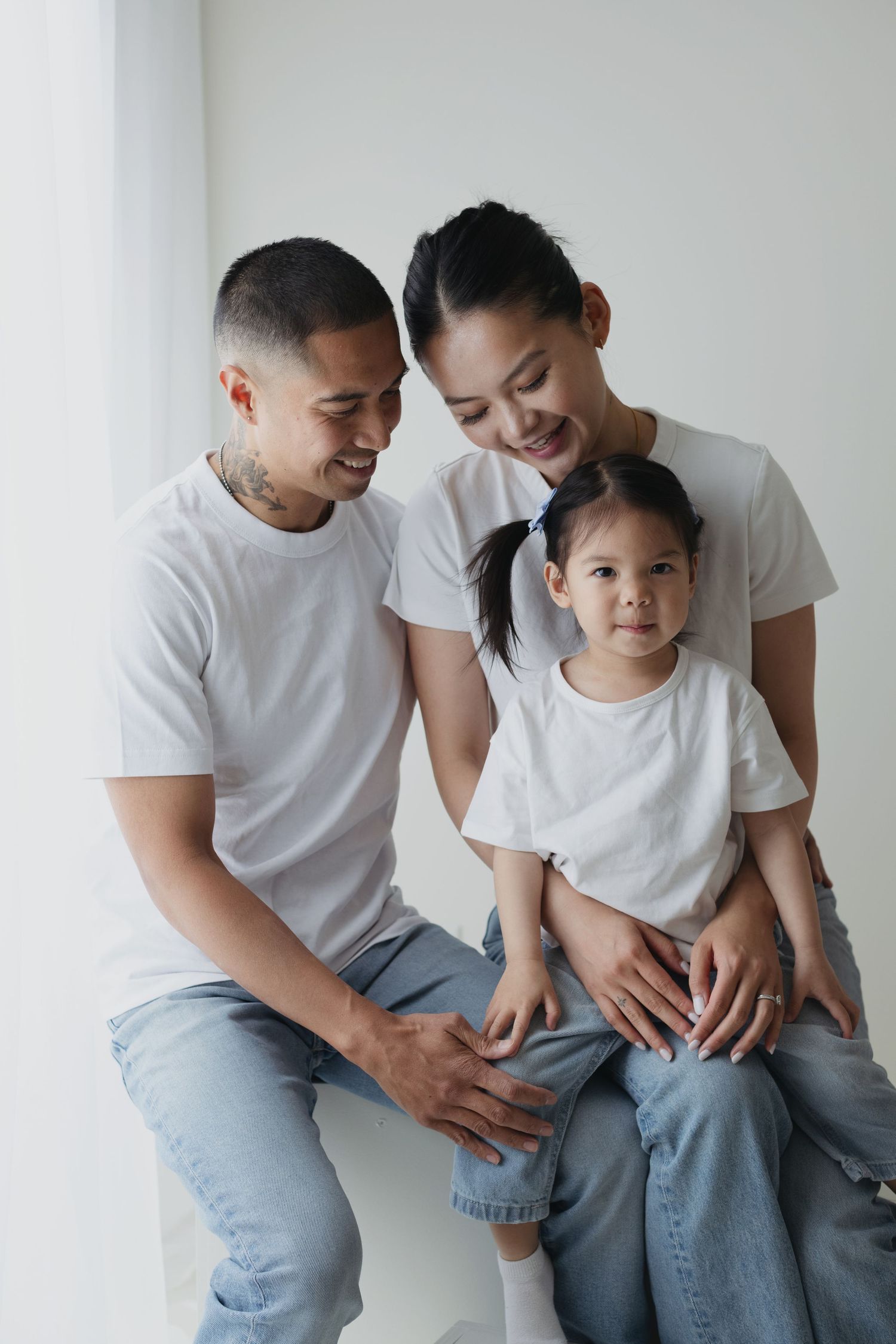 Happy Asian family of three in matching white tees and jeans posing together in a bright, minimalist studio setting.