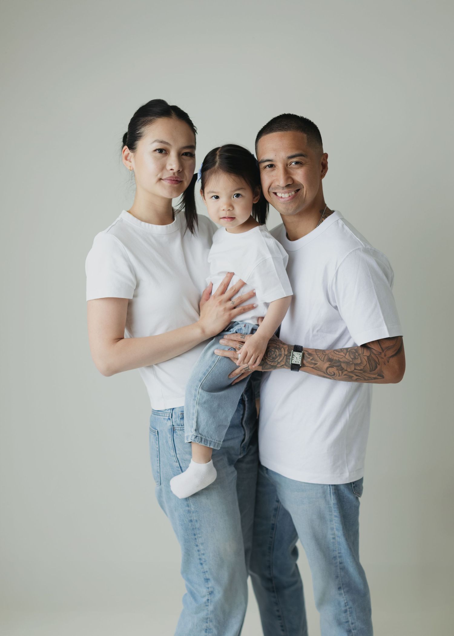 A happy family of three in matching white tees and jeans pose for a studio portrait, parents holding young daughter.