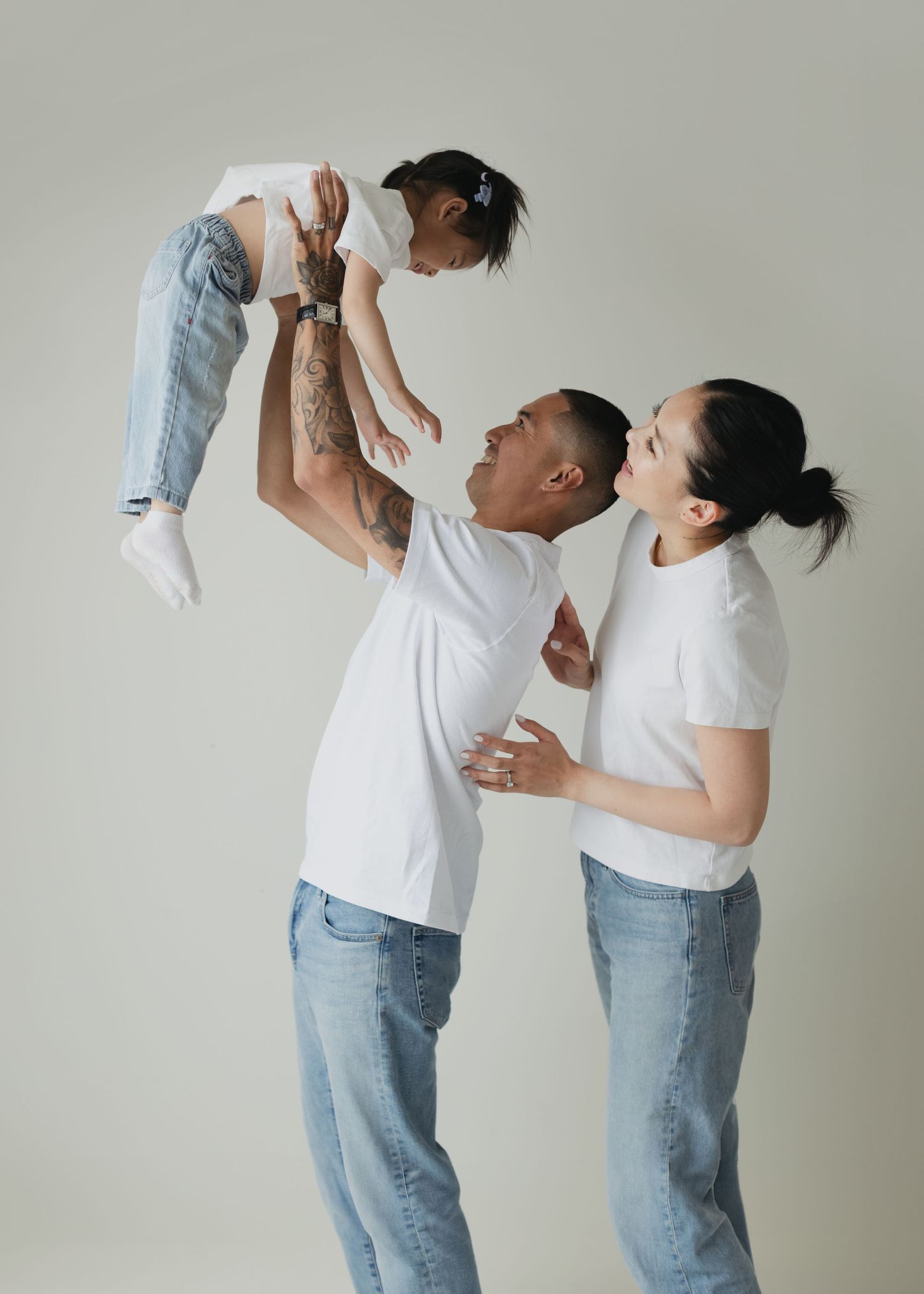 A joyful family in matching white tees and jeans, with a father lifting a baby while mother watches and smiles.