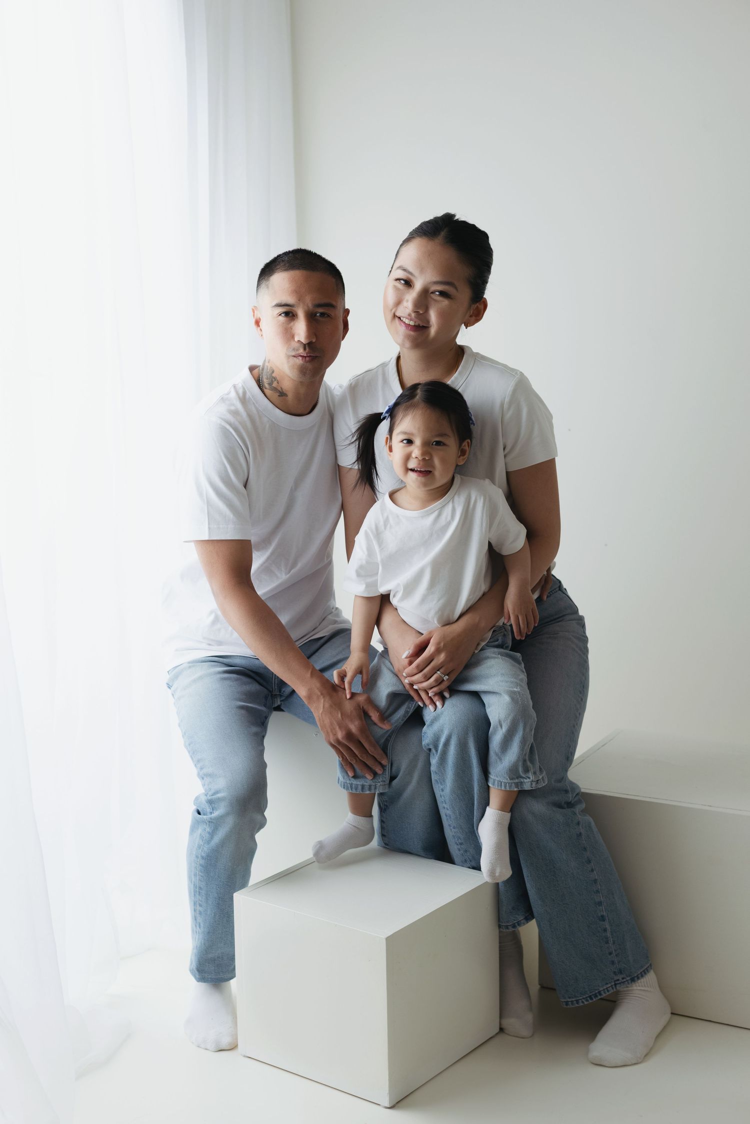 Asian family of three in matching white tees and jeans posing together on white cube in bright studio.
