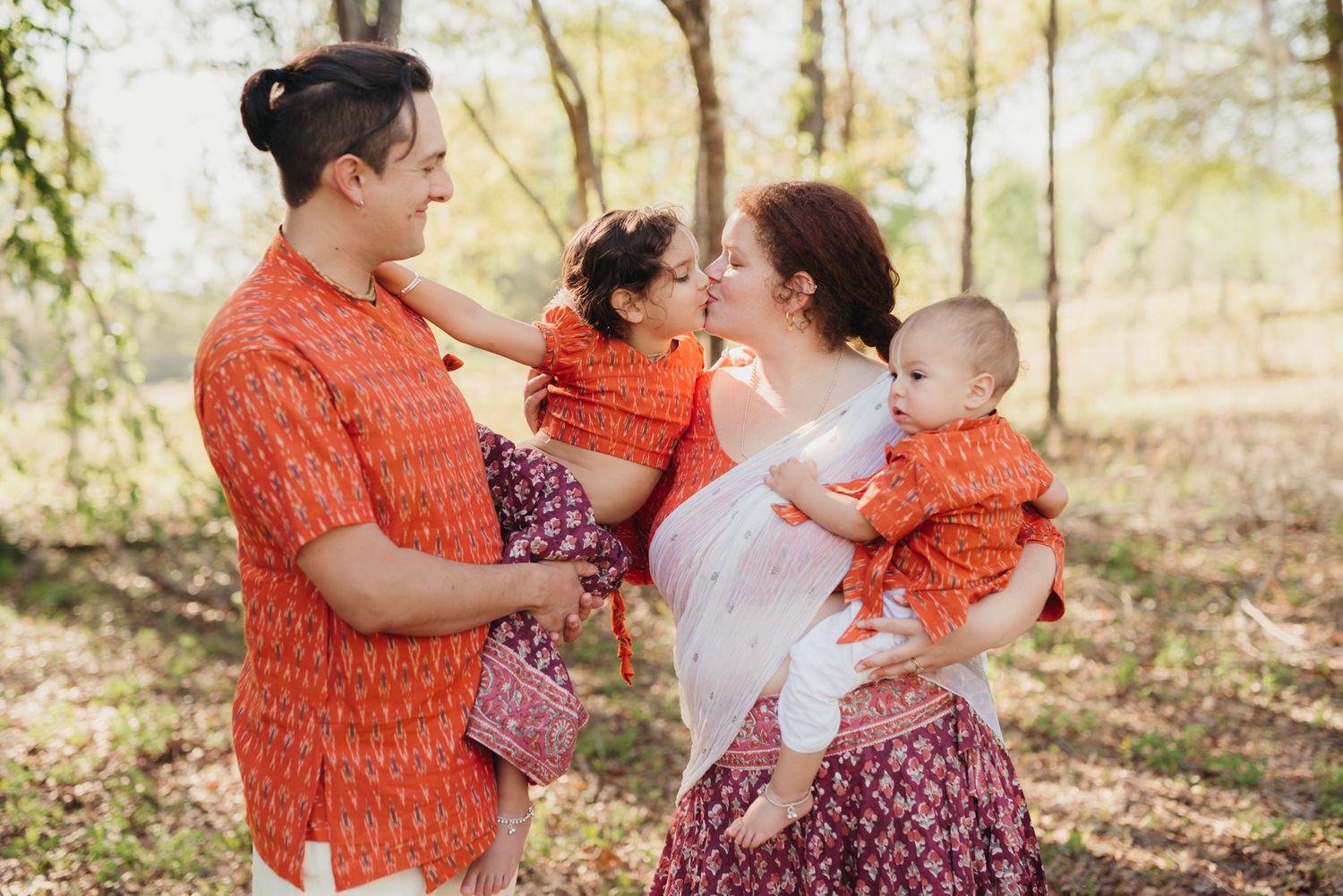 Family of four in matching orange patterned outfits stands outdoors in a sunlit woodland, parents holding two young children.