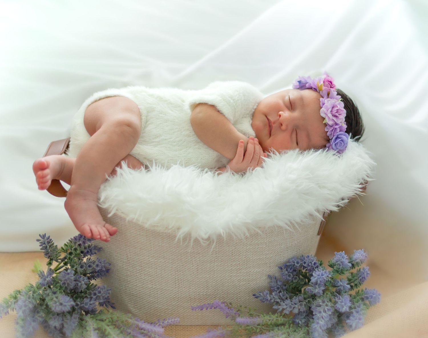Sleeping newborn baby in white fur basket with purple flower headband, surrounded by lavender blooms.