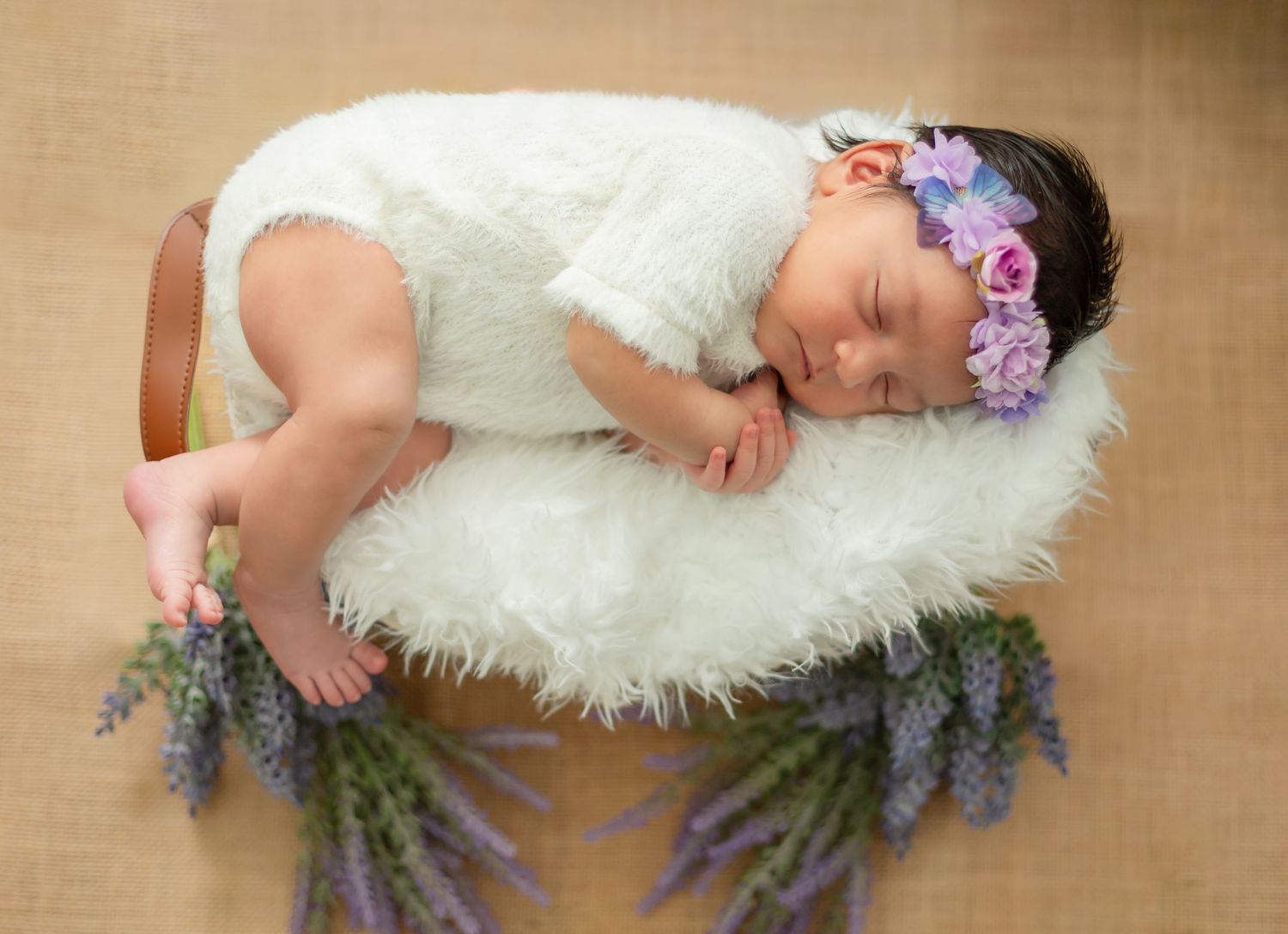 Sleeping newborn baby in white romper on fluffy rug with purple floral headband and lavender bouquets.