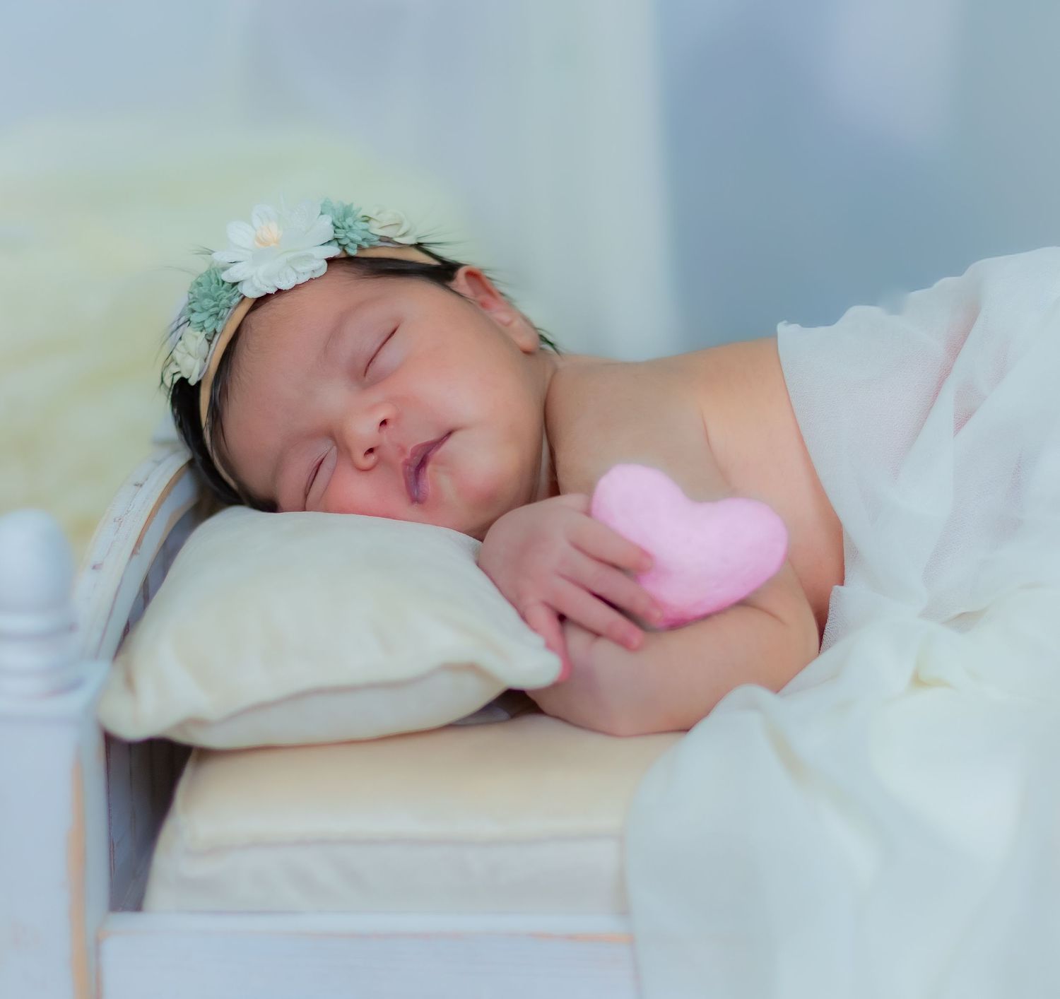 Sleeping newborn baby girl wearing floral headband, holding pink heart toy on white pillow.