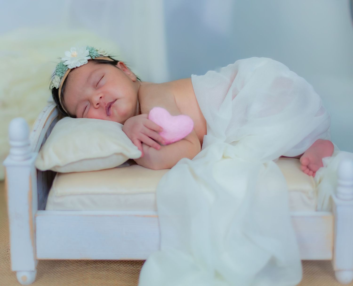 Peaceful newborn baby girl in white tulle wrap holding a pink heart, sleeping on a tiny white bed.