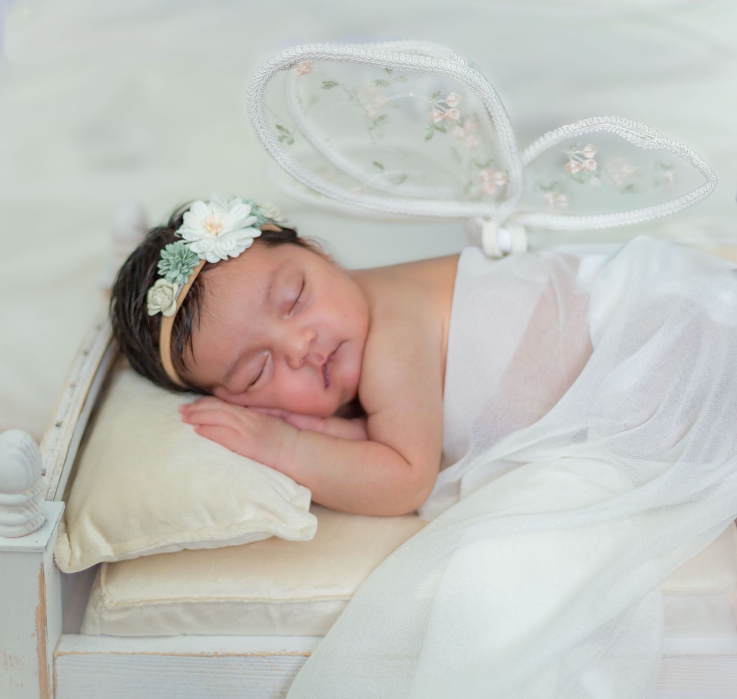 Sleeping newborn baby girl wearing white bunny ears headband and white tulle wrap on a miniature bed.