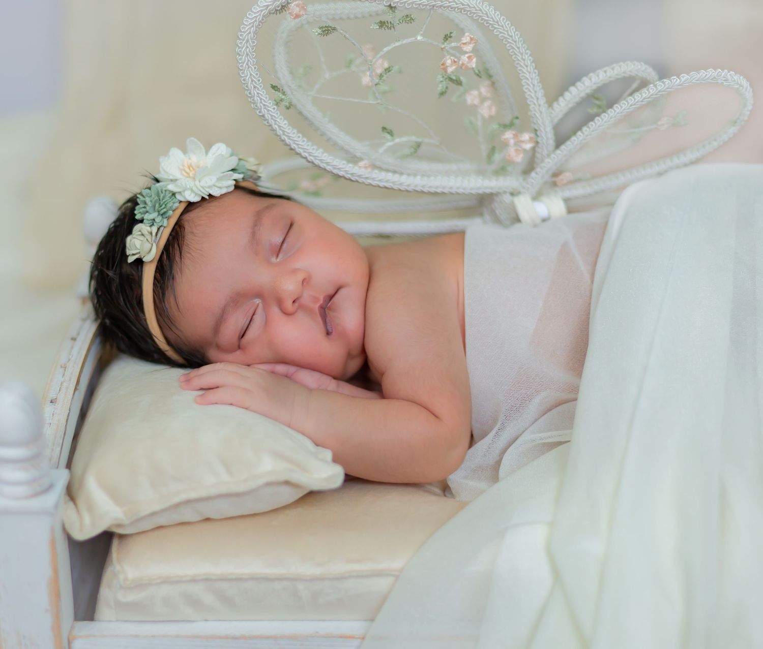 Peaceful newborn baby sleeping on a pillow wearing a floral headband in a white decorative bed.