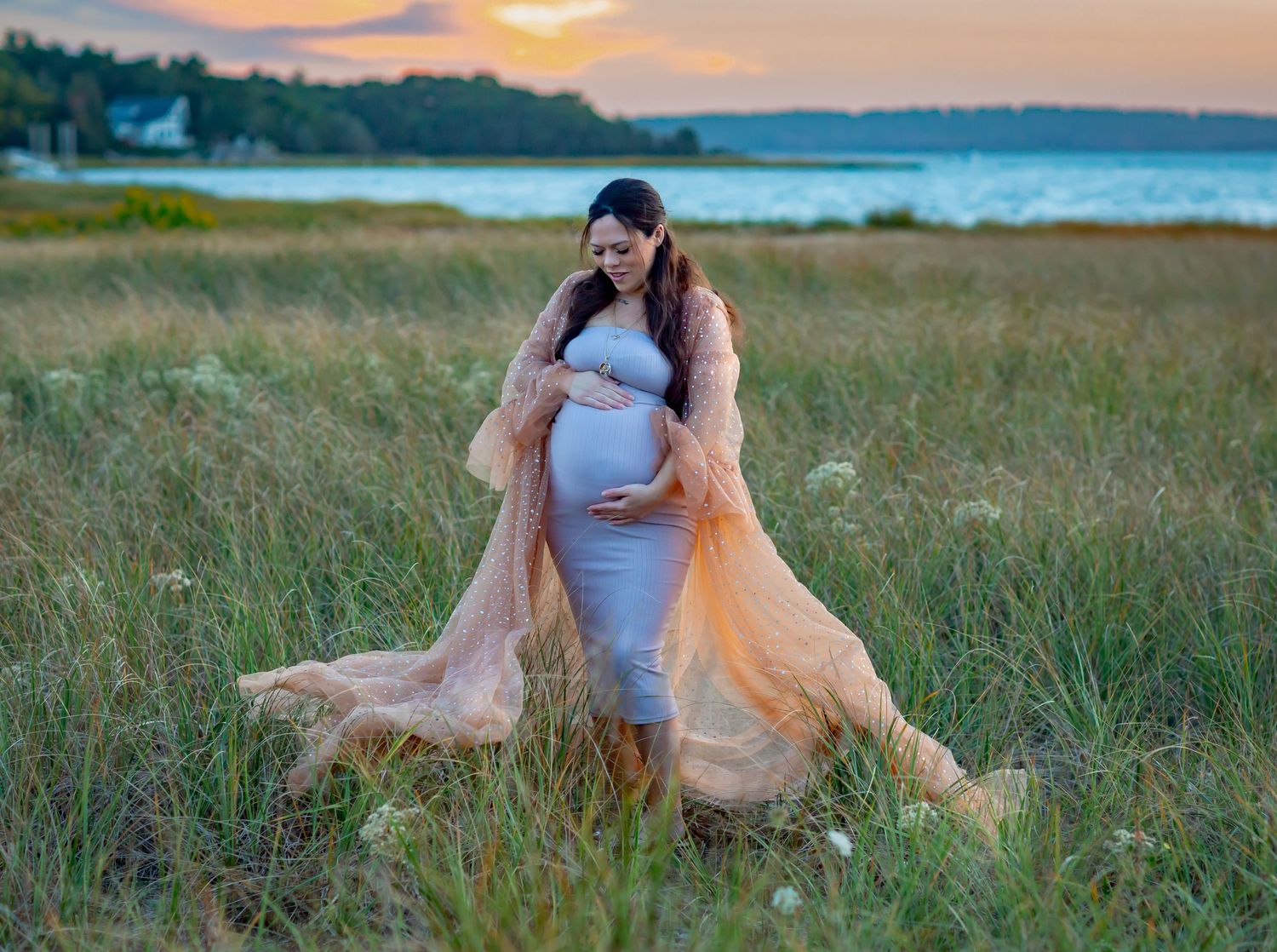 Pregnant woman in gray dress and peach robe poses in golden field by water at sunset.
