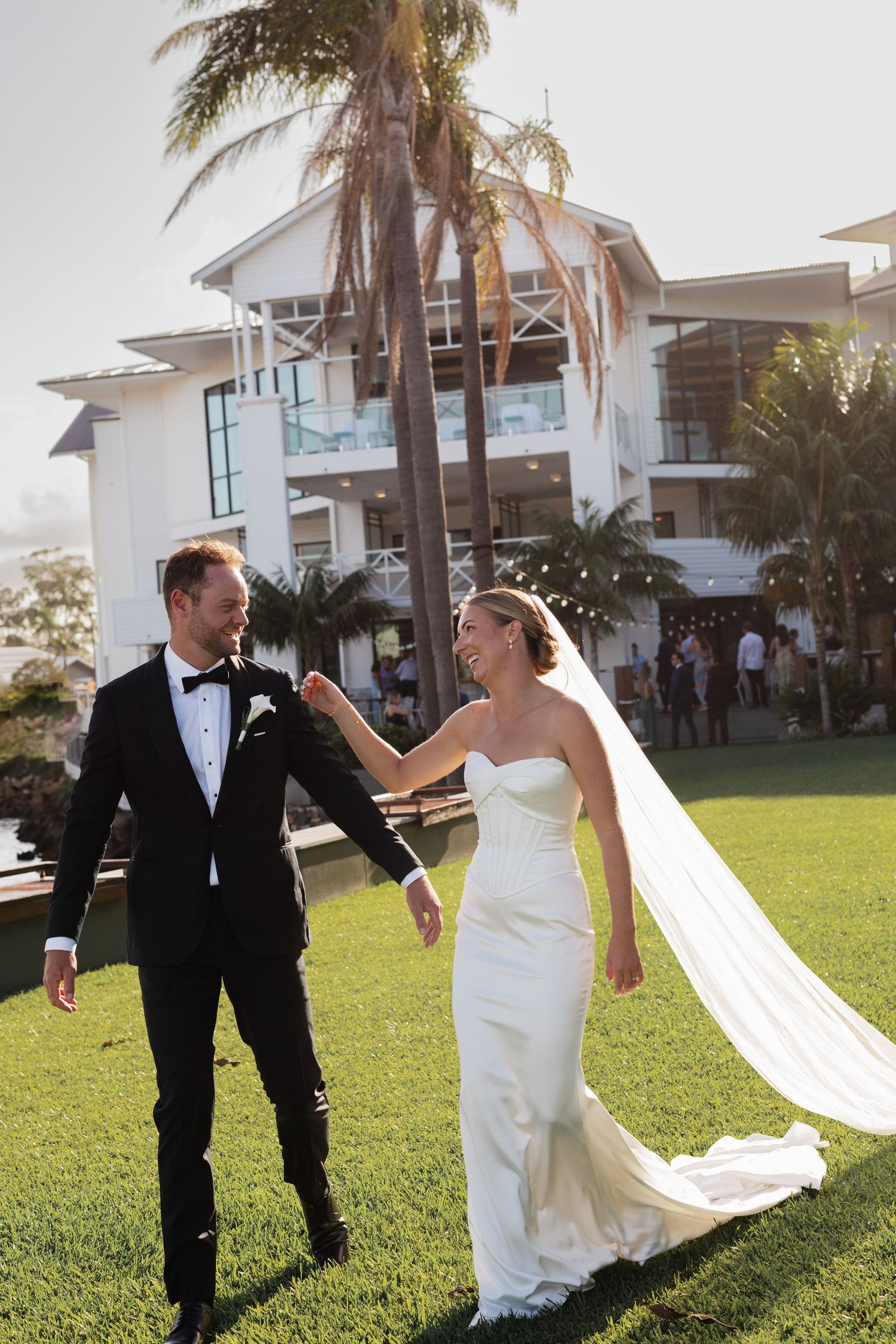 A bride and groom share a romantic kiss on a waterfront lawn with a palm tree and white venue building behind them.