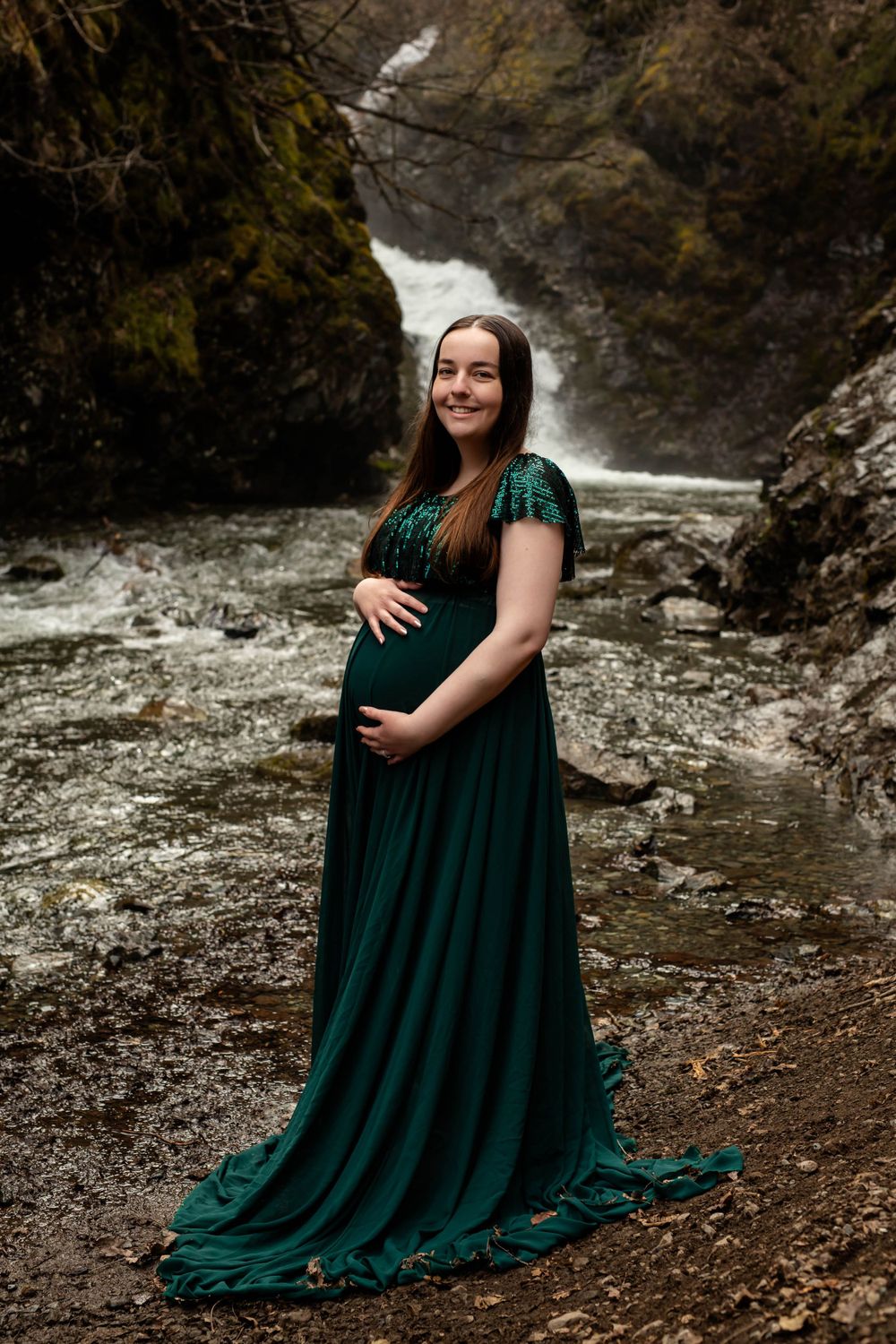 Maternity portrait in emerald green dress against dramatic waterfall backdrop.