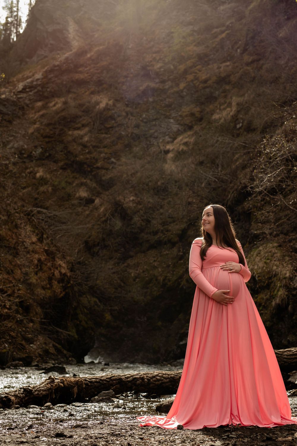 Maternity photo featuring coral floor-length dress against rocky coastal backdrop.