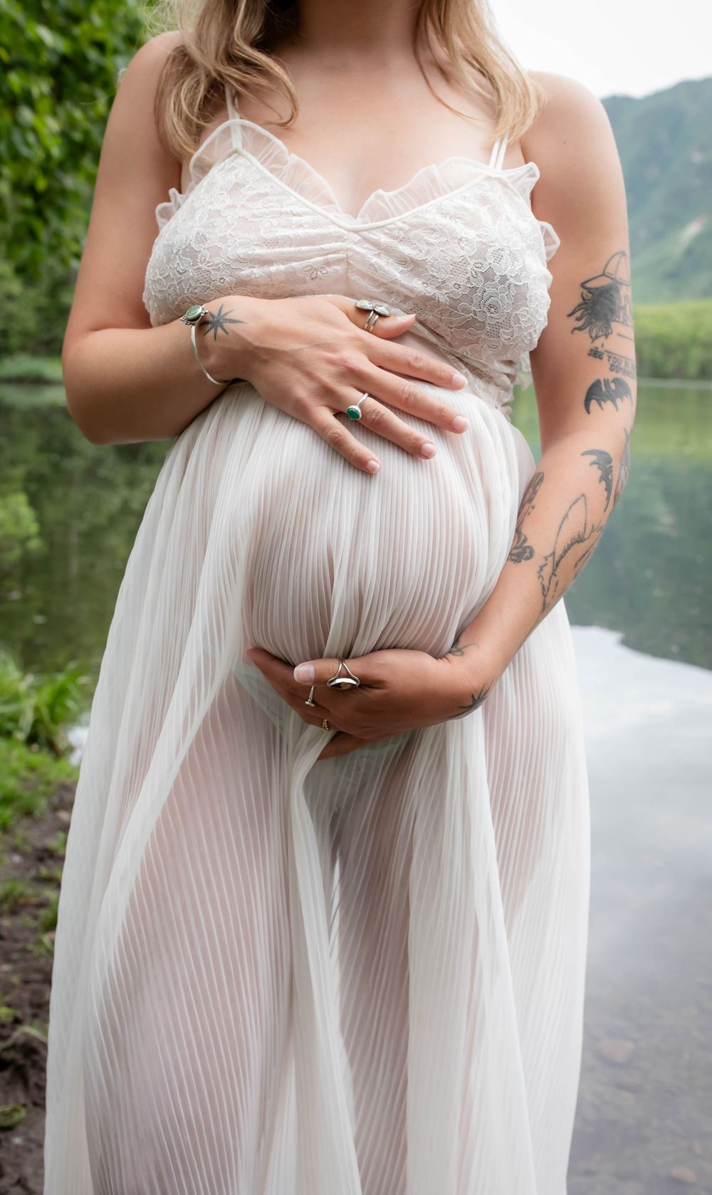 A maternity photo showing a white lace dress and visible tattoos against a lakeside mountain backdrop.