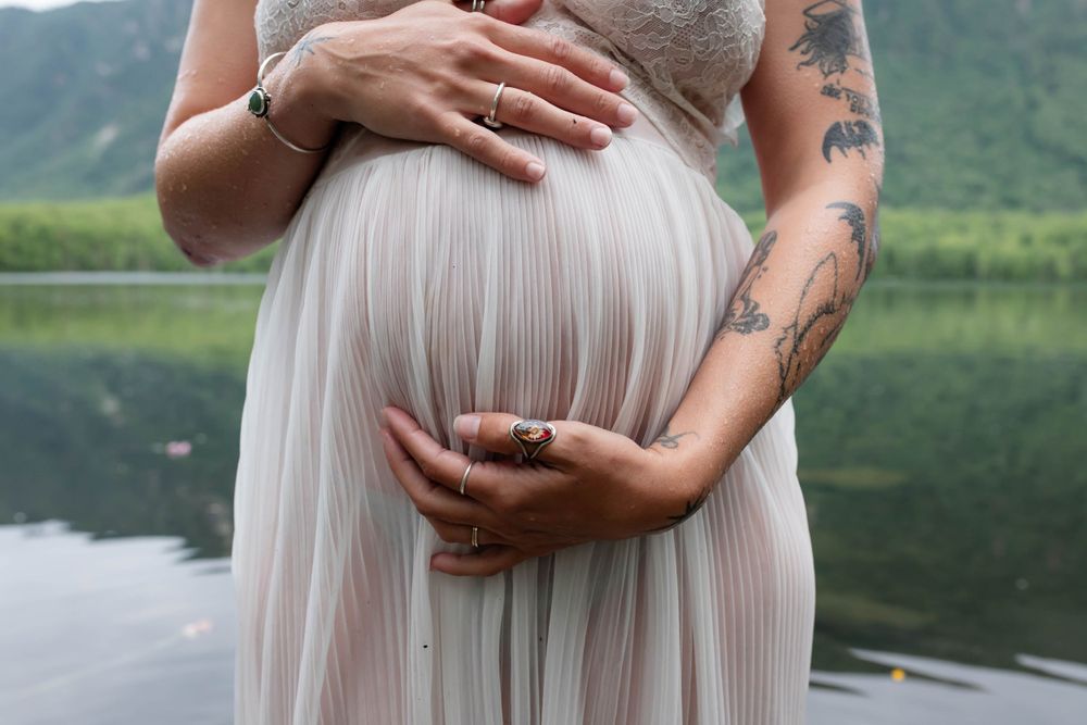 Close-up of hands cradling pregnant belly in pleated white dress by mountain lake.