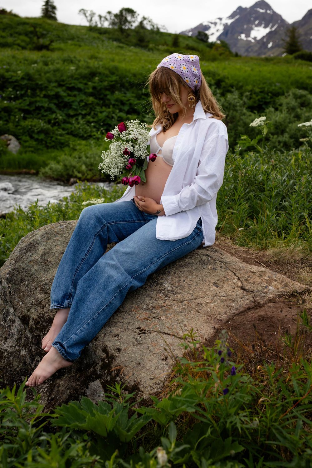 Person in casual attire sits on rock by mountain stream holding wildflower bouquet.