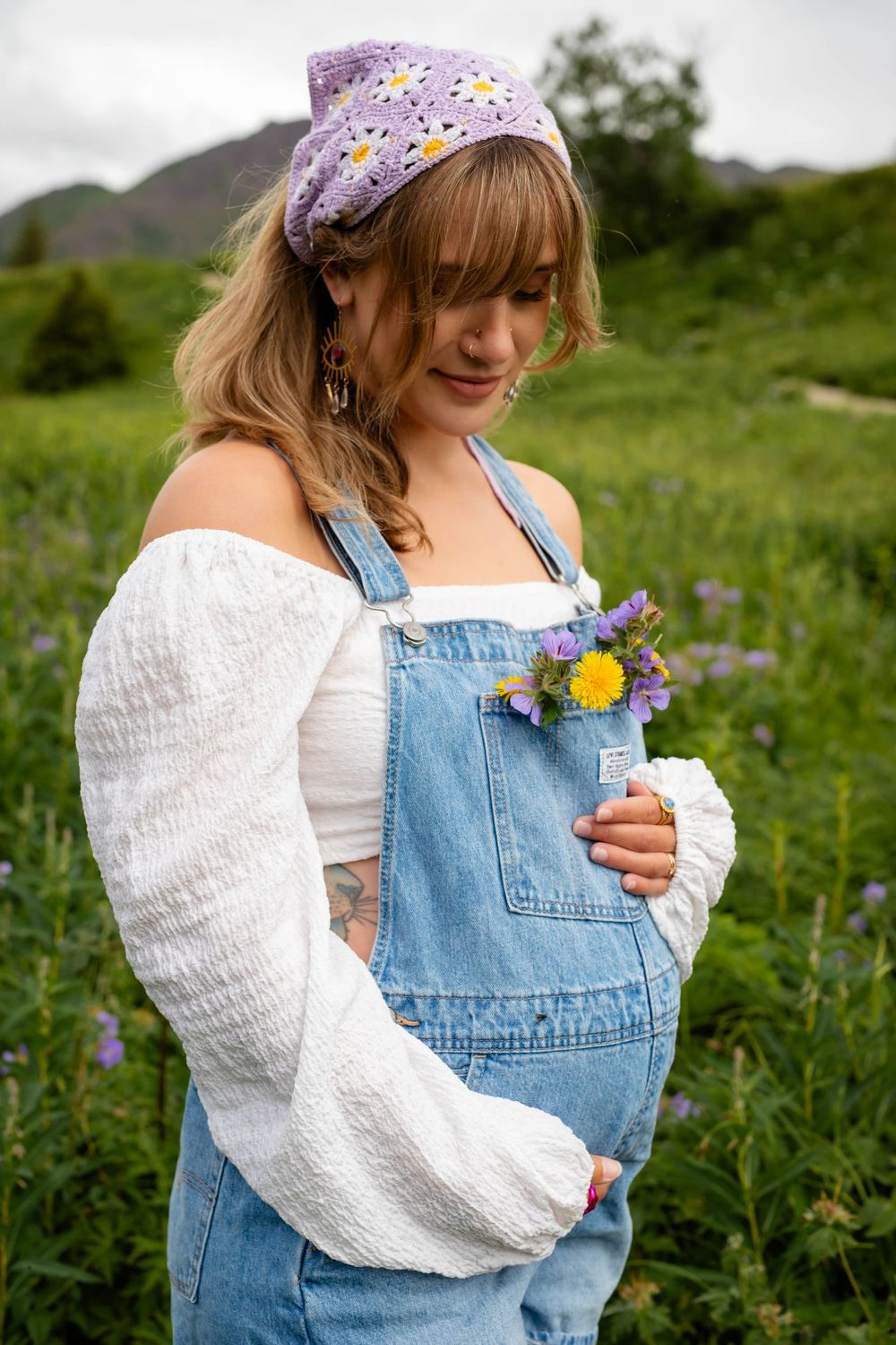 Person in overalls and floral bandana holds wildflowers in mountain meadow.