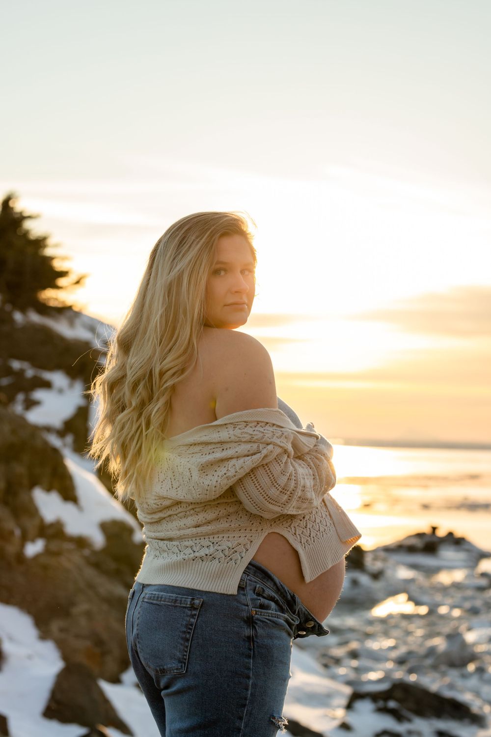 Maternity sunset portrait with sweater and jeans against snowy coastal backdrop.