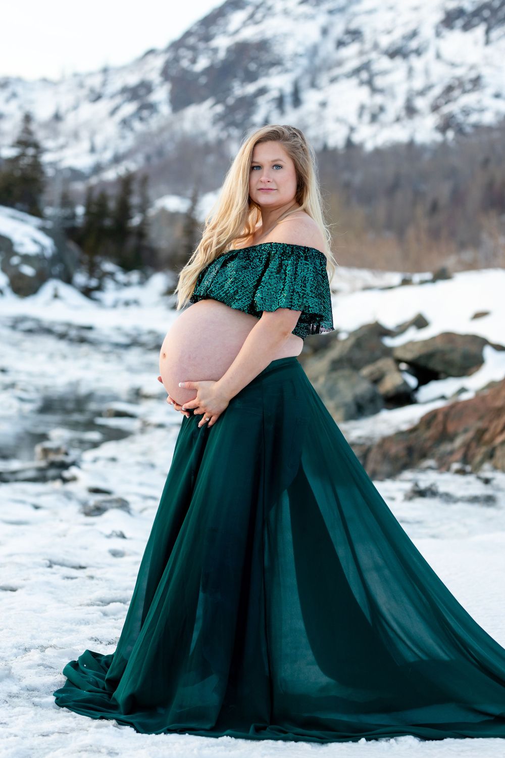 Pregnant woman in flowing emerald gown poses in snowy mountain landscape.