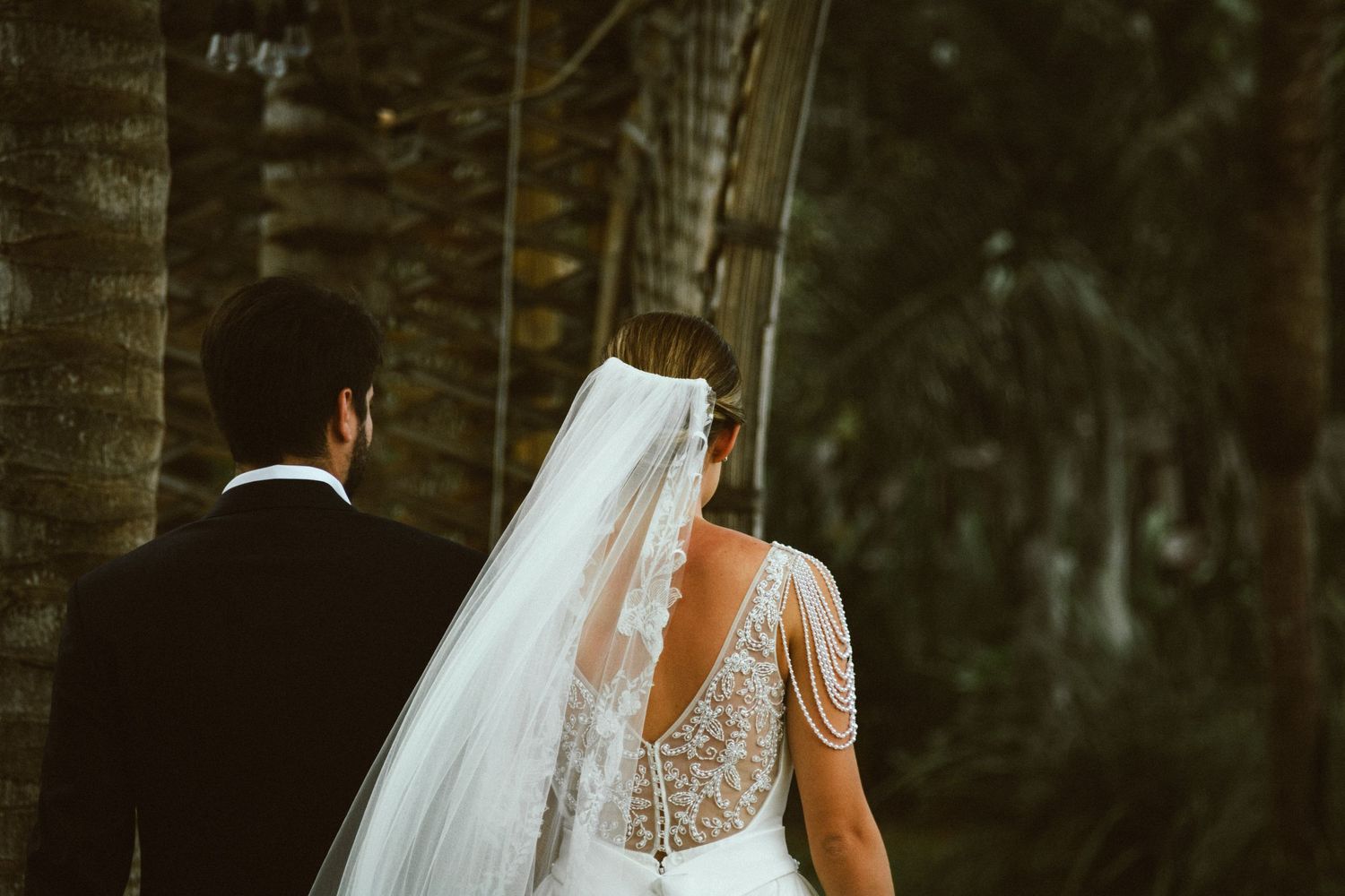 Bride and groom walking among palm trees, showcasing elegant lace wedding dress with veil.