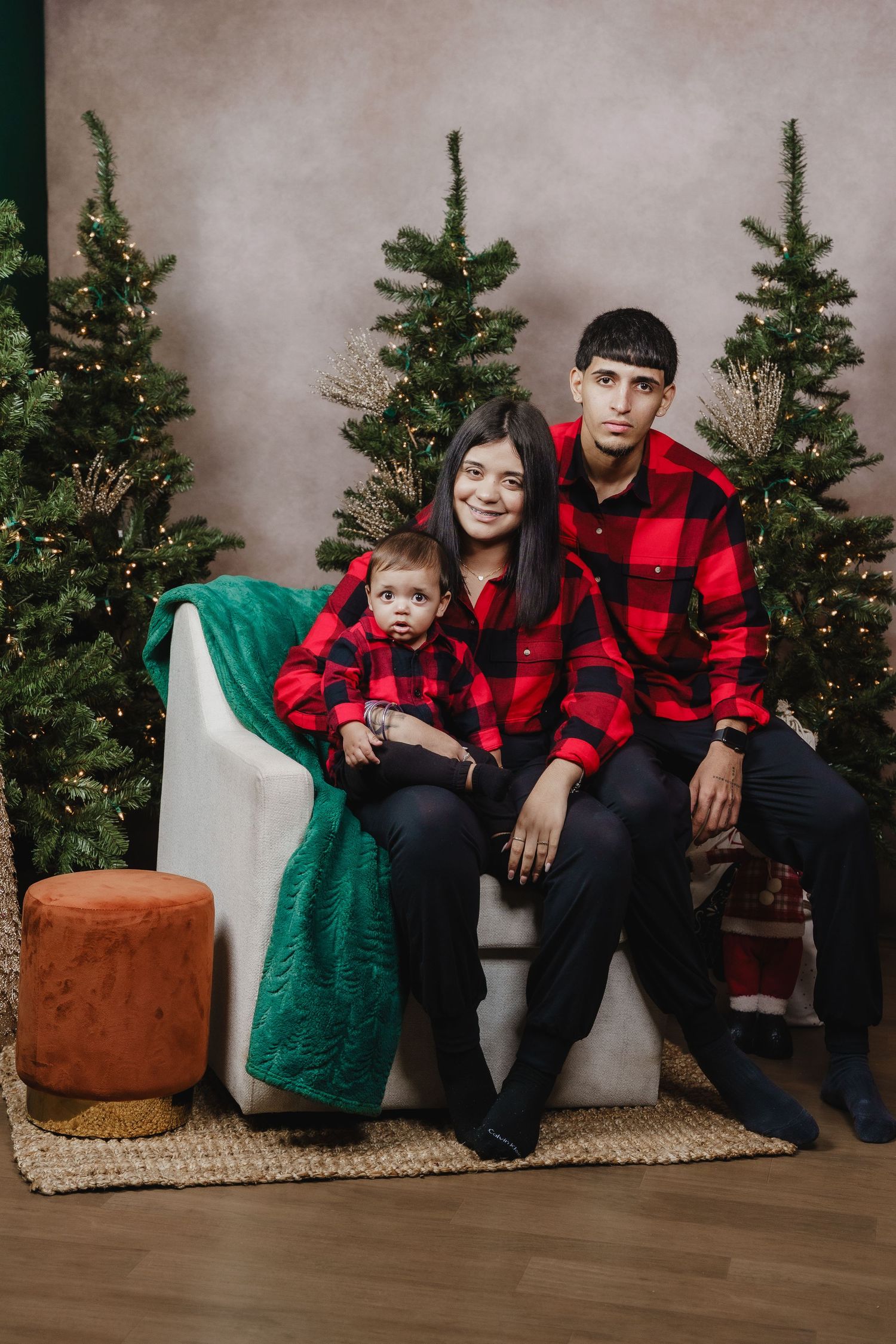 Family in coordinating red buffalo plaid outfits poses for holiday portrait with Christmas trees.