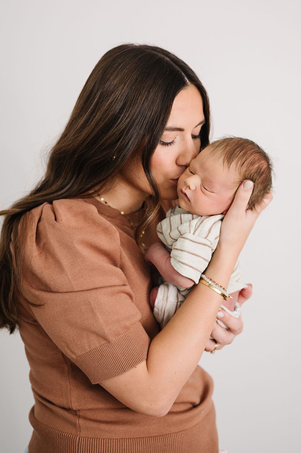 An intimate portrait of a mother in brown sweater tenderly kissing her sleeping newborn.