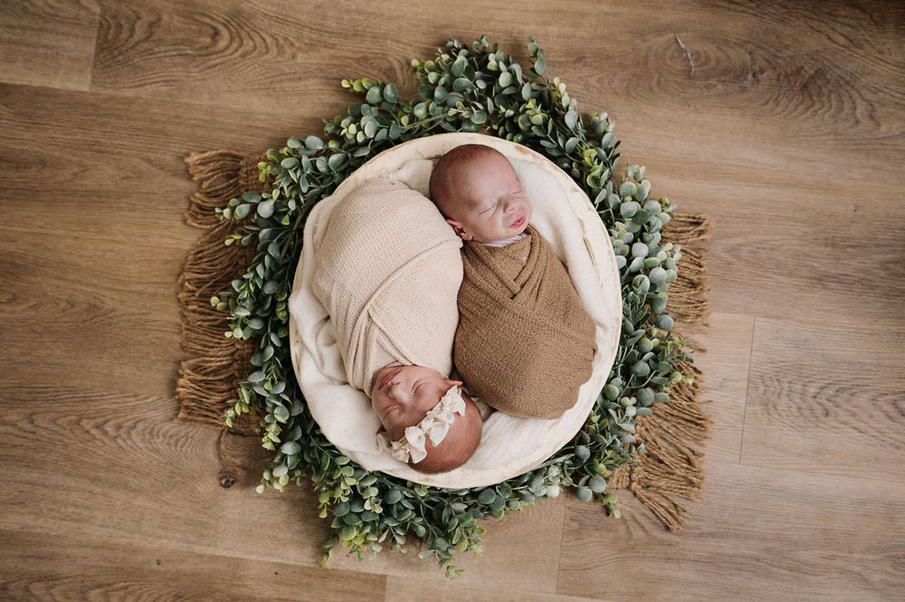 Sleeping newborn in brown wrap nestled in white cushion surrounded by greenery wreath.