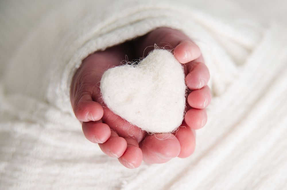 Baby feet holding white heart shape against white blanket background.