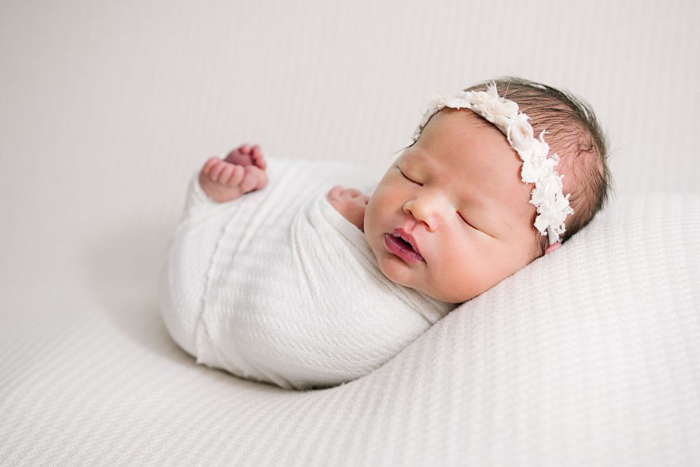 Sleeping newborn in white wrap with lace headband on white textured surface.