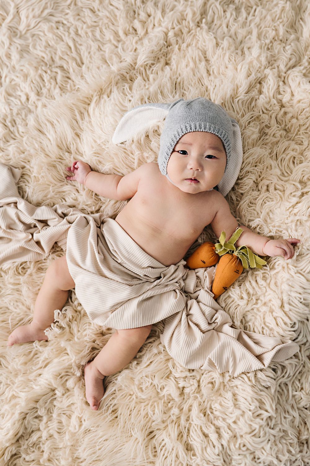 Baby in grey hat lying on cream rug with decorative carrots.
