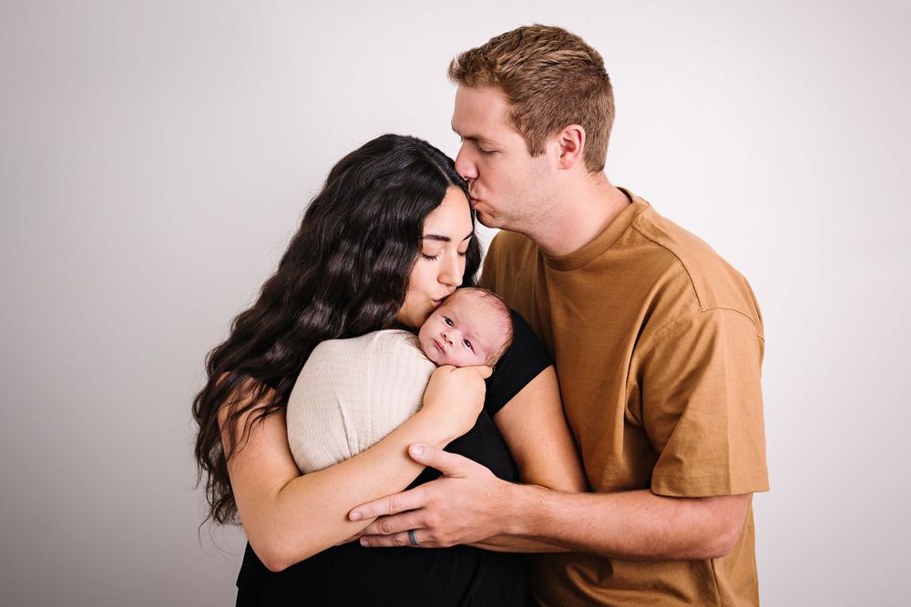 Intimate family moment with couple embracing swaddled baby against light background.