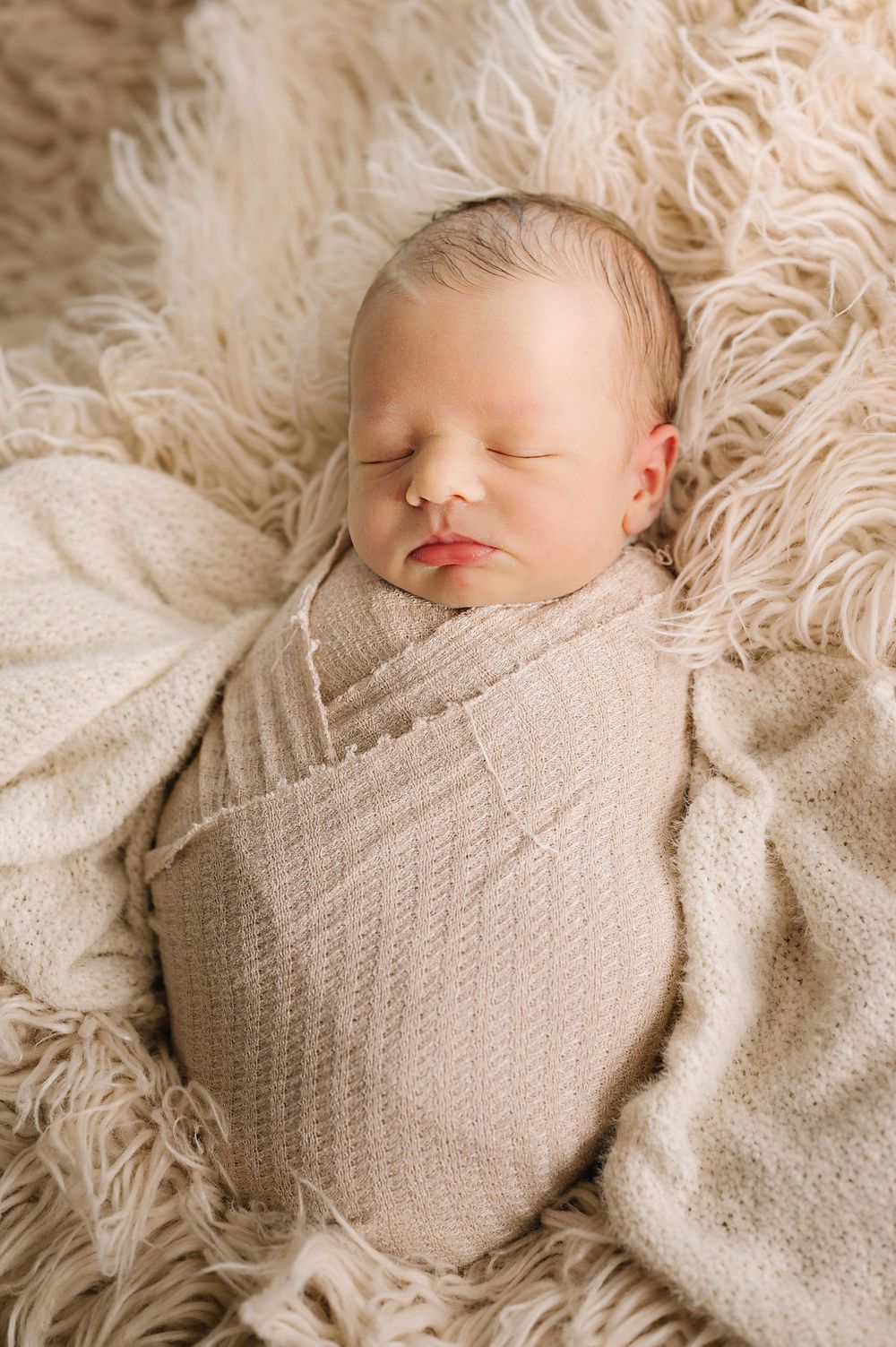 Newborn baby sleeping peacefully while swaddled in beige wrap on fluffy cream-colored fur.