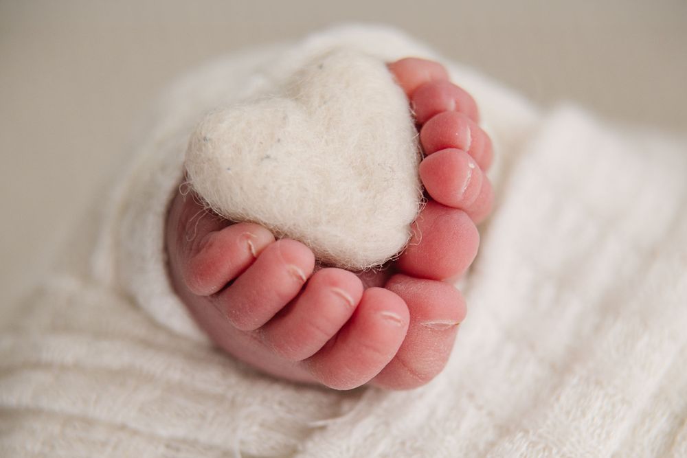 A tiny baby foot holding a white heart-shaped object on a white blanket.