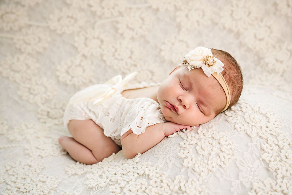 Newborn baby wearing white lace outfit and delicate headband sleeping peacefully on textured white blanket.